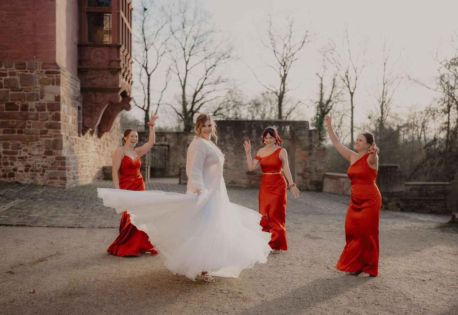 Wedding party in red dresses and white gown posed against rustic stone building exterior.