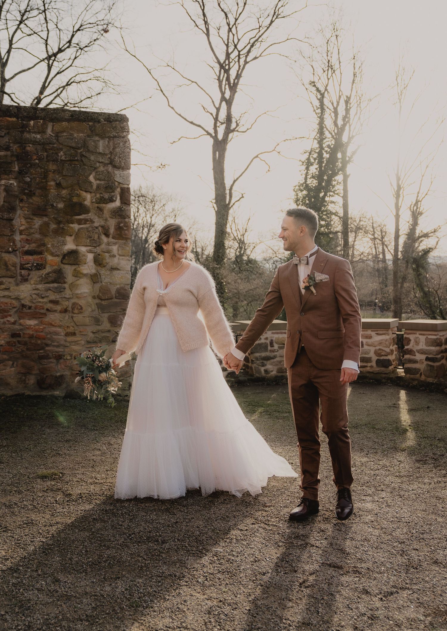 Candid wedding couple shares playful moment together on outdoor path by stone wall at sunset.