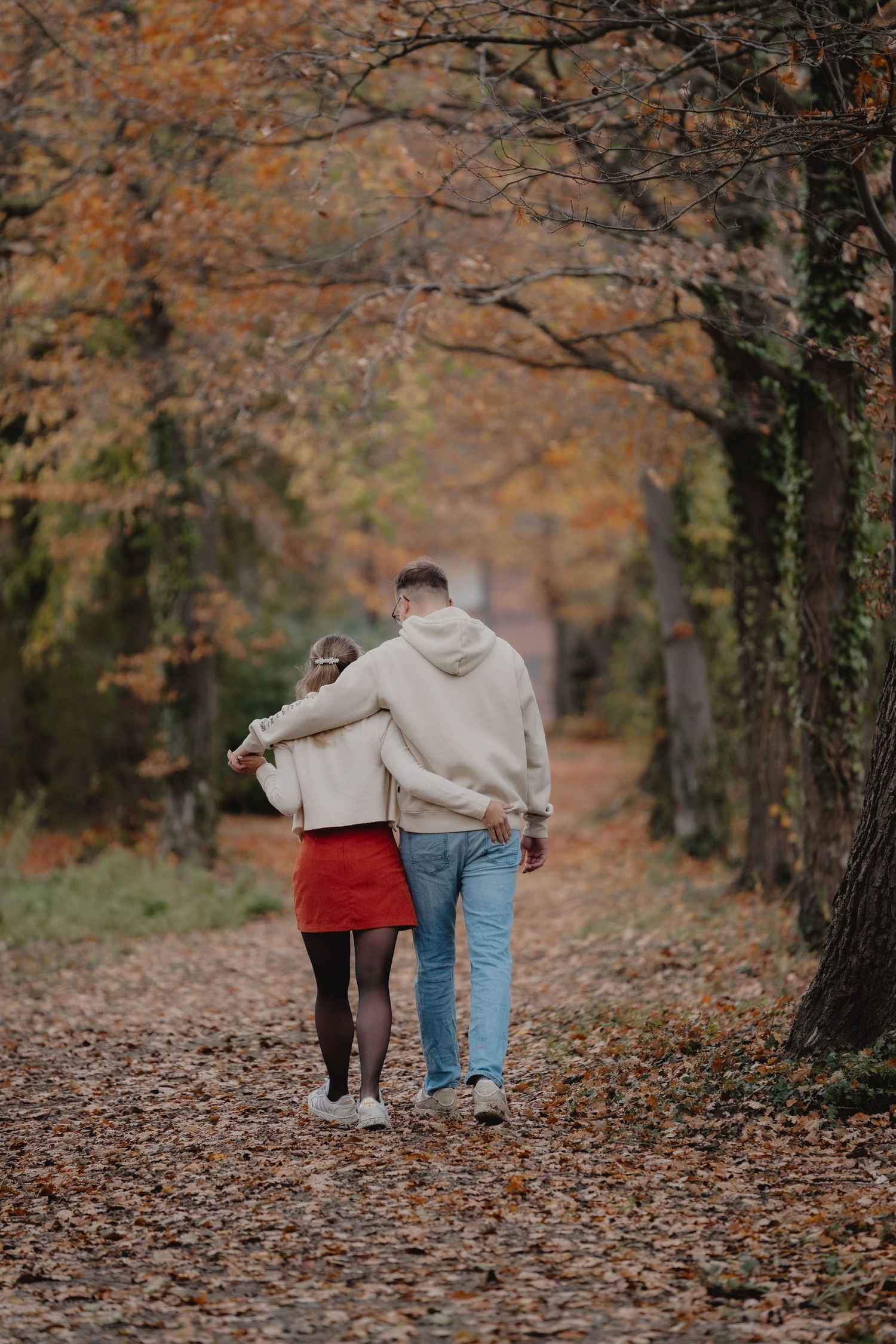 A couple walks away hand in hand down a leaf-covered autumn path.