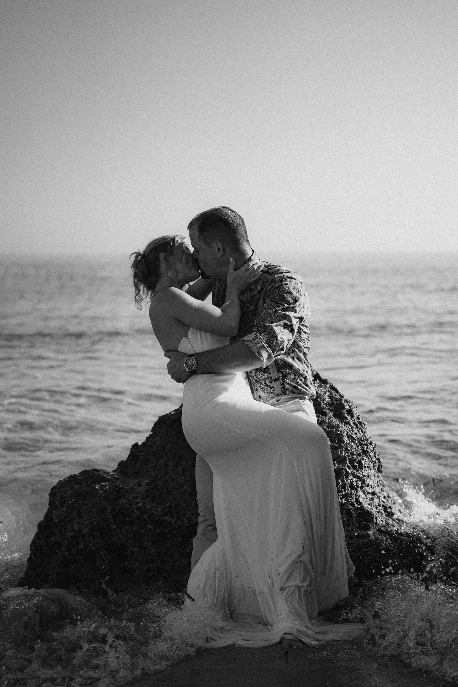 A romantic black and white photo of a couple embracing on ocean rocks at sunset.