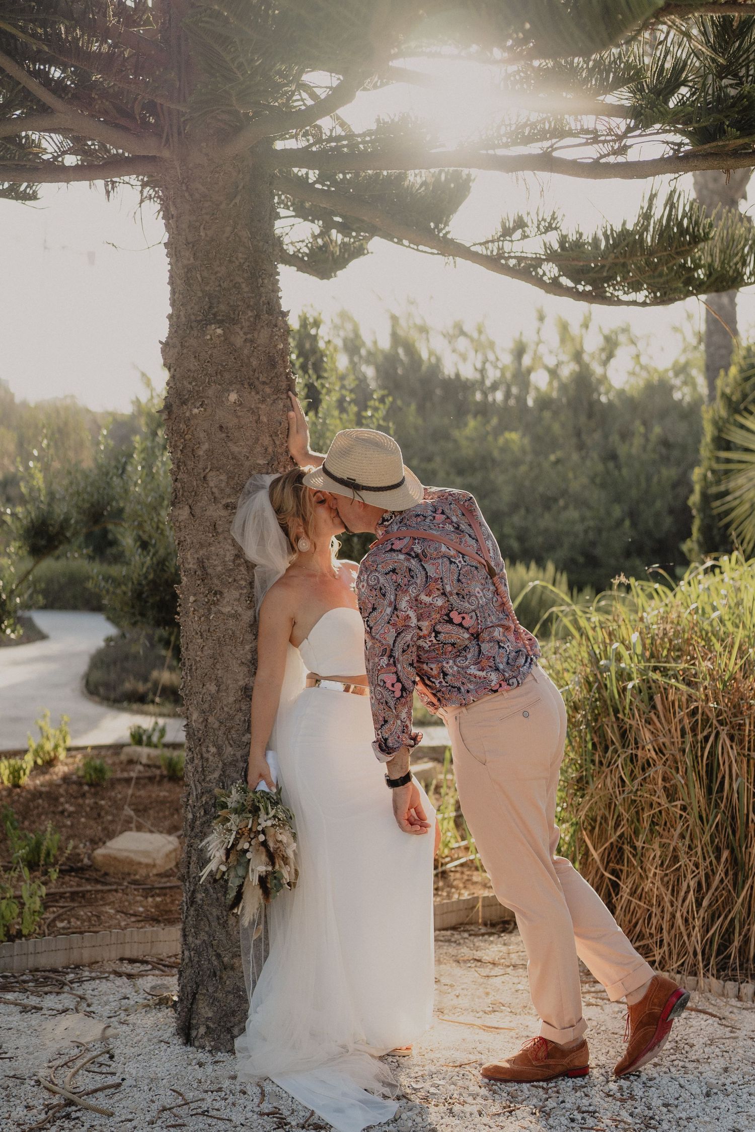 Sunset wedding photo of a couple in white dress and floral shirt walking together among palm trees.
