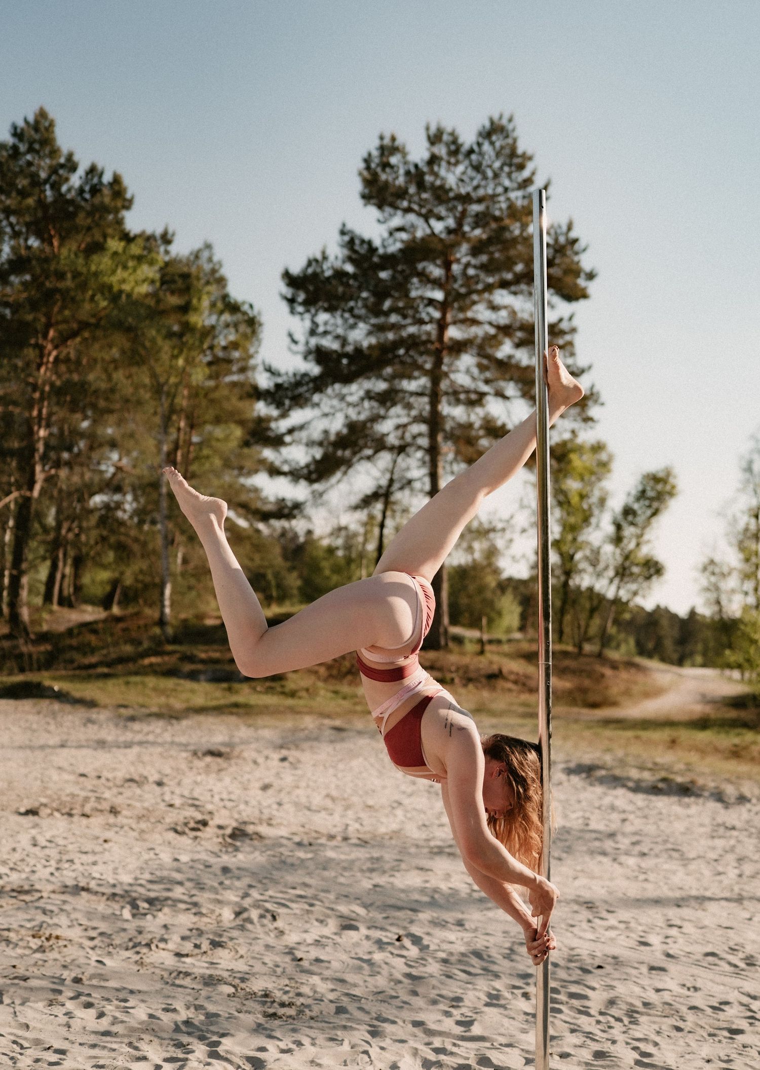 Woman performing an impressive pole dance move outdoors on a sandy beach surrounded by tall pine trees.