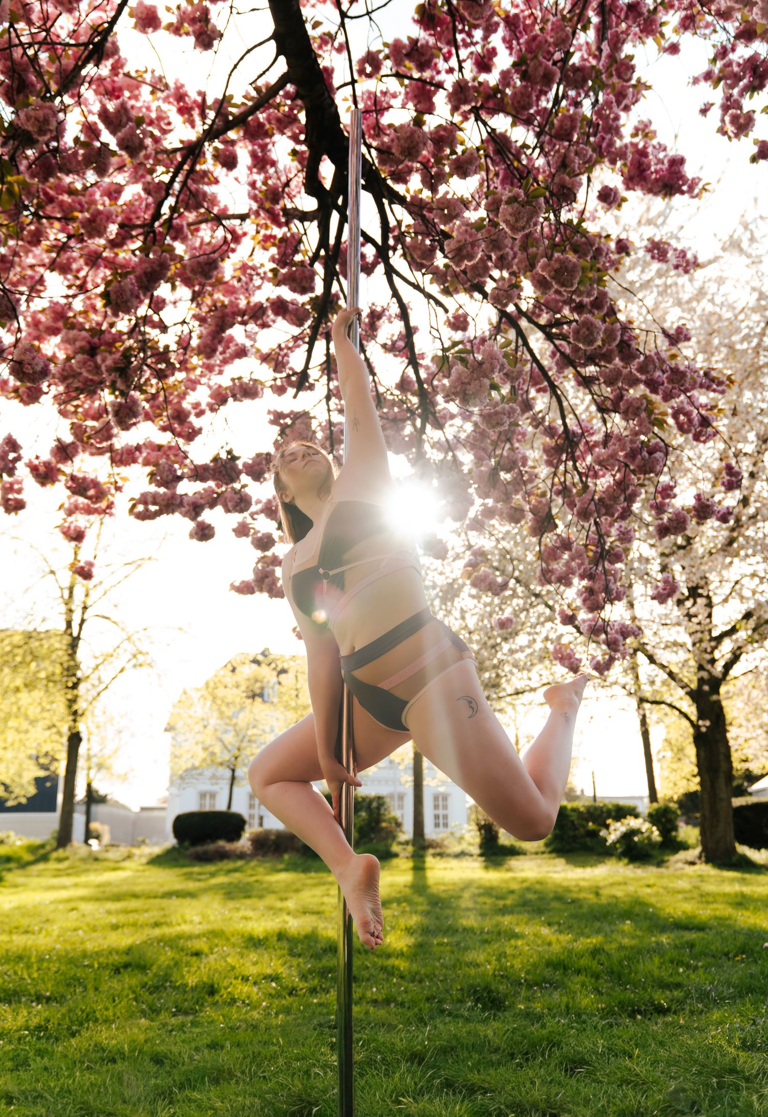 Woman pole dancing outdoors under blooming pink cherry blossom trees in green bikini on sunny spring day.