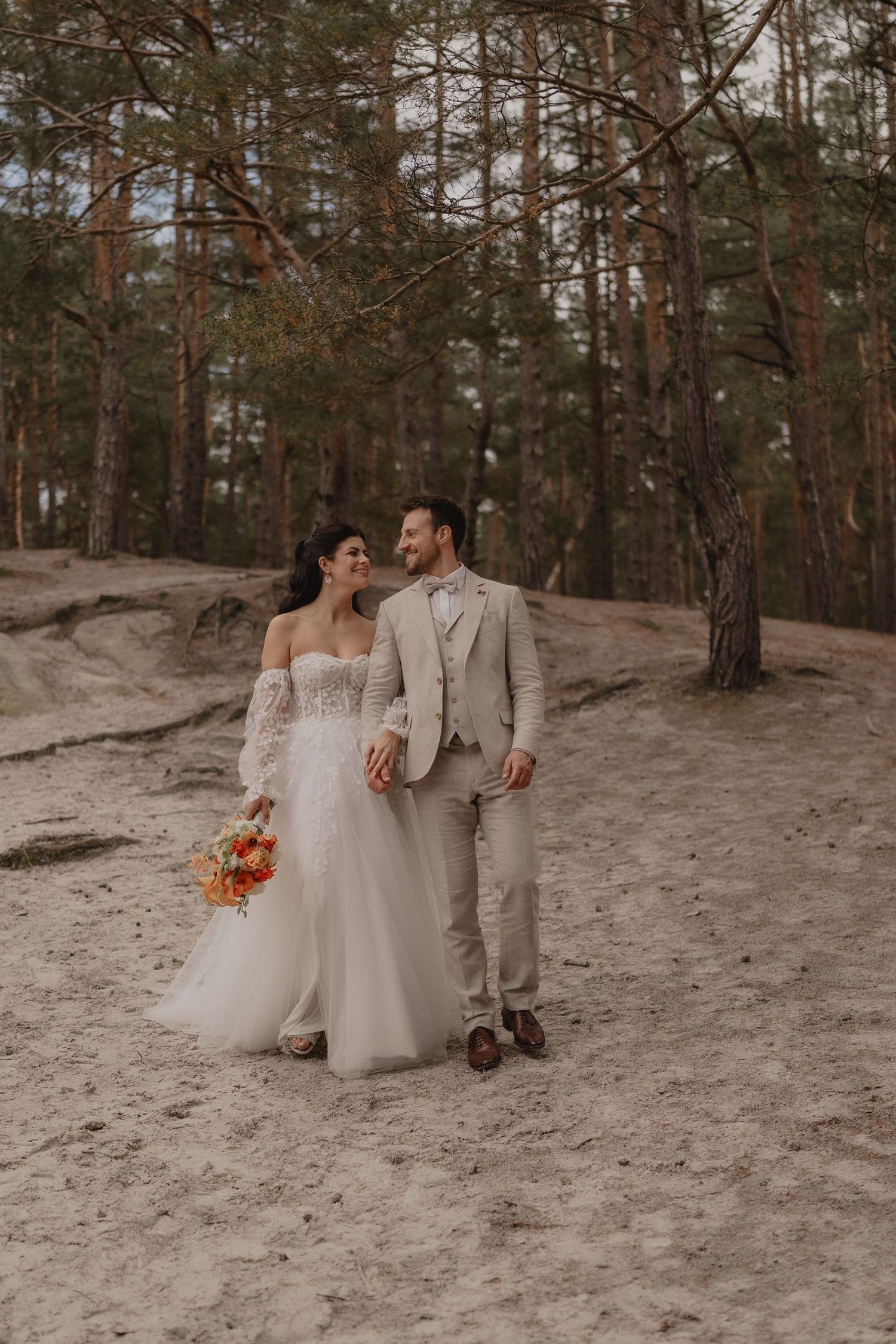 A couple in wedding attire walk together through a pine forest holding orange flowers on a sandy path.
