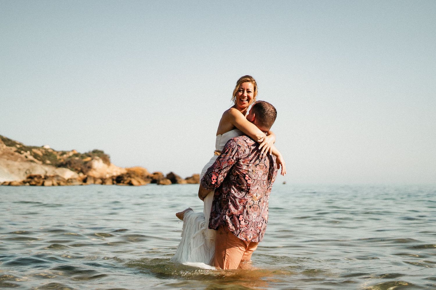 A couple embraces in knee-deep ocean water with rocky coastline visible in the background.