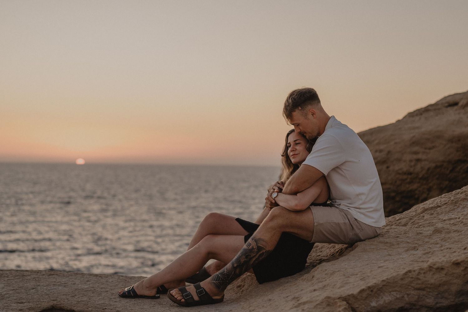 Couple embracing at sunset on rocky beach, golden hour ocean backdrop.