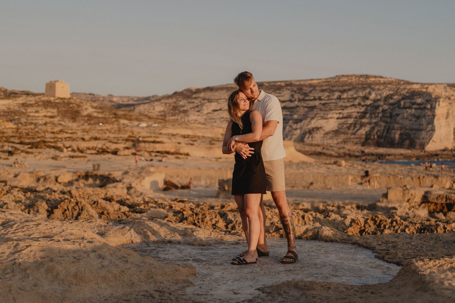 Couple embracing and kissing at golden hour in a rocky desert landscape with sandy cliffs in the background.