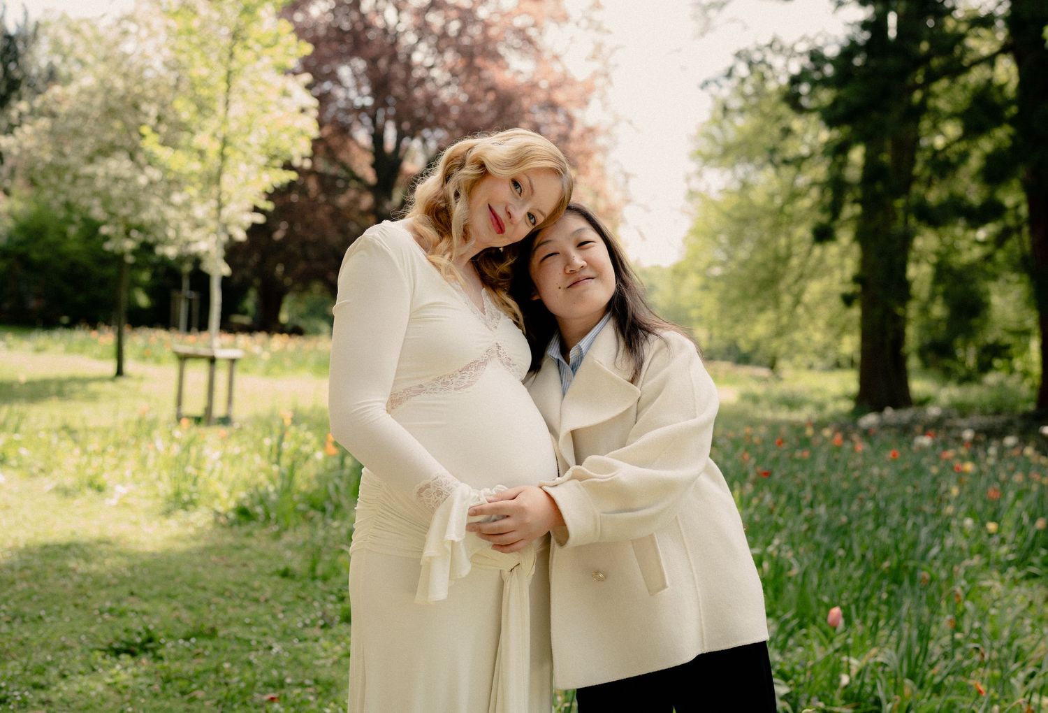 Pregnant woman in white dress embraced by friend in lush garden with spring flowers.