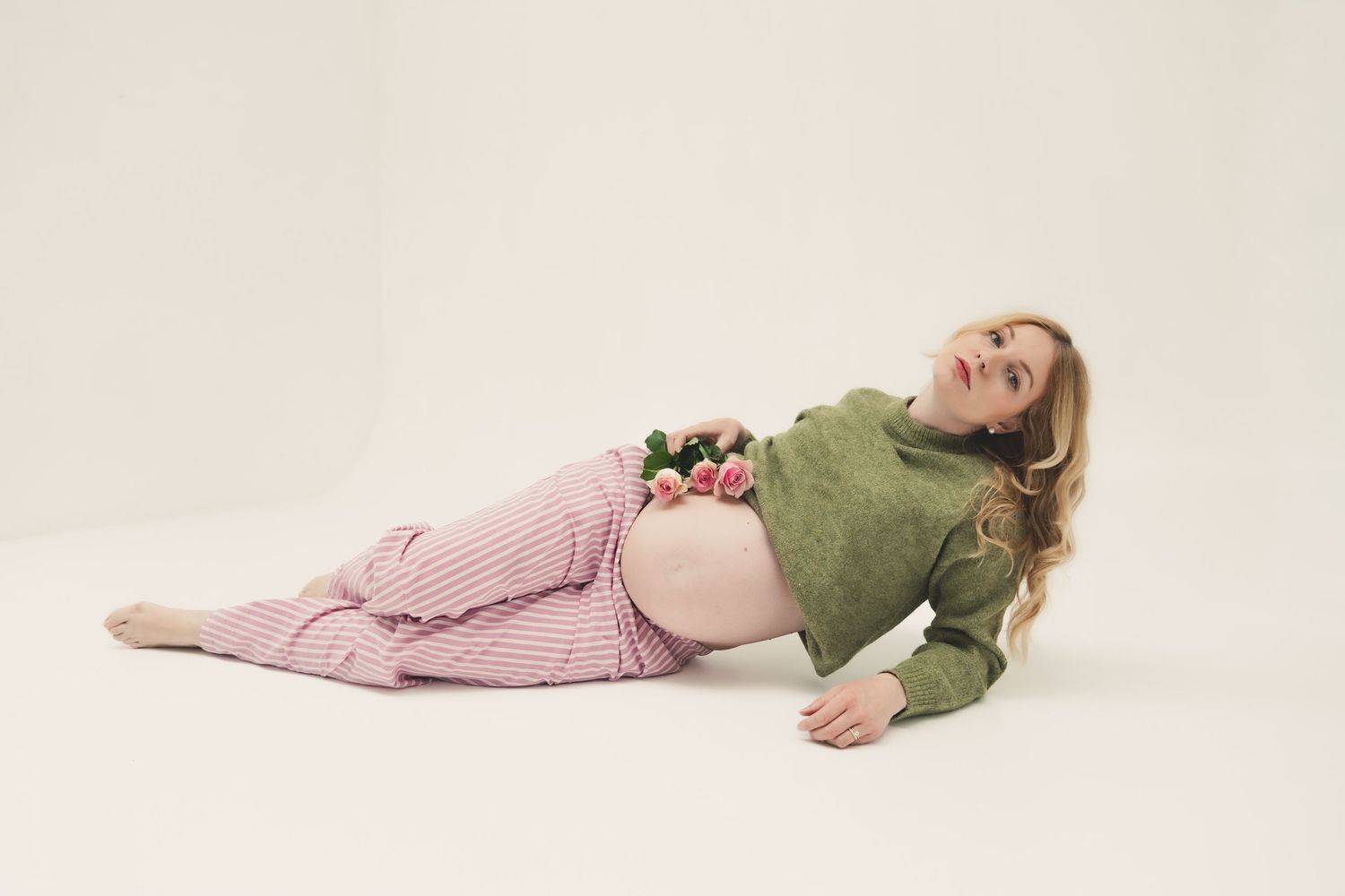 Pregnant woman lying on floor in green crop top and pink trousers with roses on belly against white background.