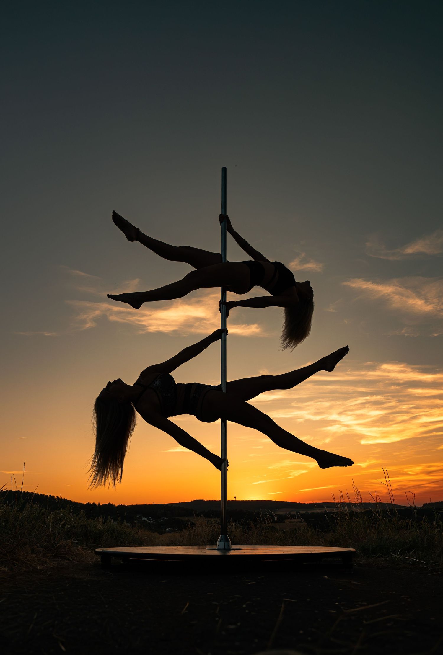 Two women pole dancing as silhouettes against a vibrant orange sunset sky outdoors.