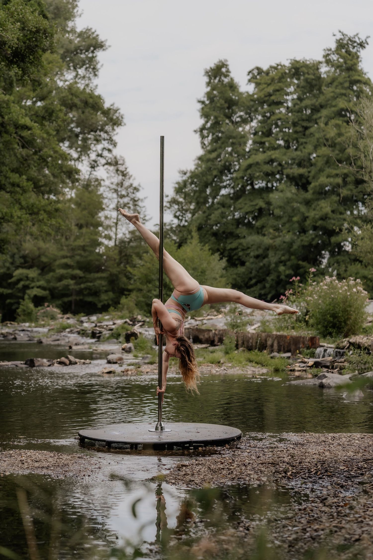 A woman performs an impressive pole dance move outdoors near a rocky stream surrounded by lush green trees.
