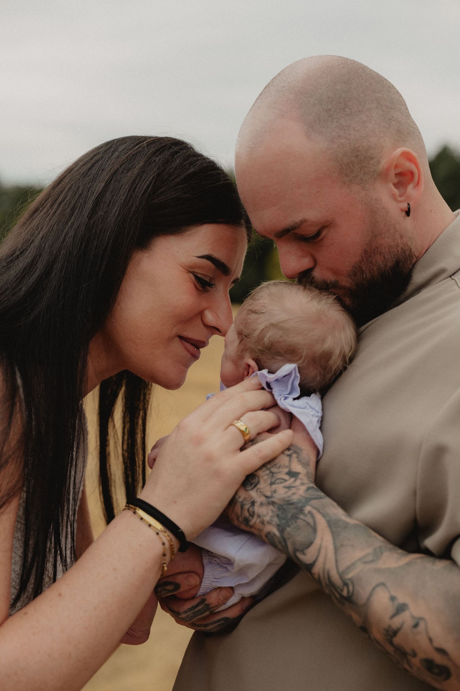 A tattooed couple tenderly kiss their newborn baby outdoors in a heartwarming family portrait.