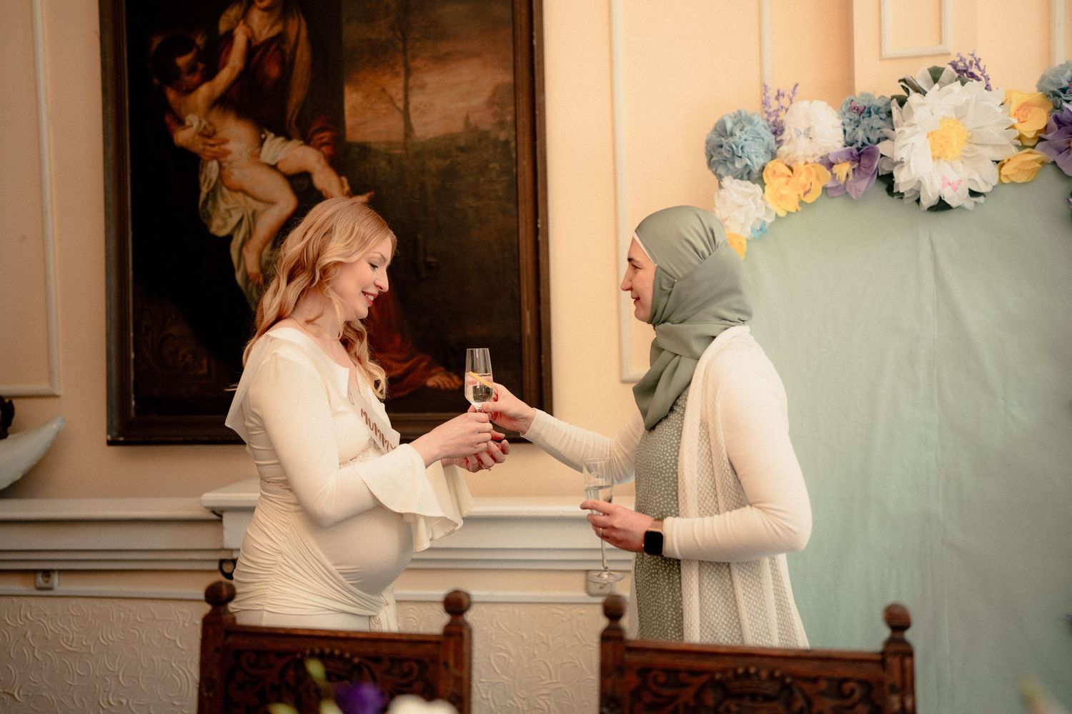 Pregnant bride in white dress toasting champagne with a woman in a sage green hijab.