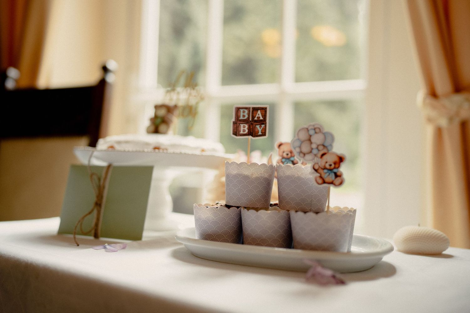 Baby shower table display with chocolate letter blocks spelling BABY, cupcakes, and a teddy bear topper on white plate.