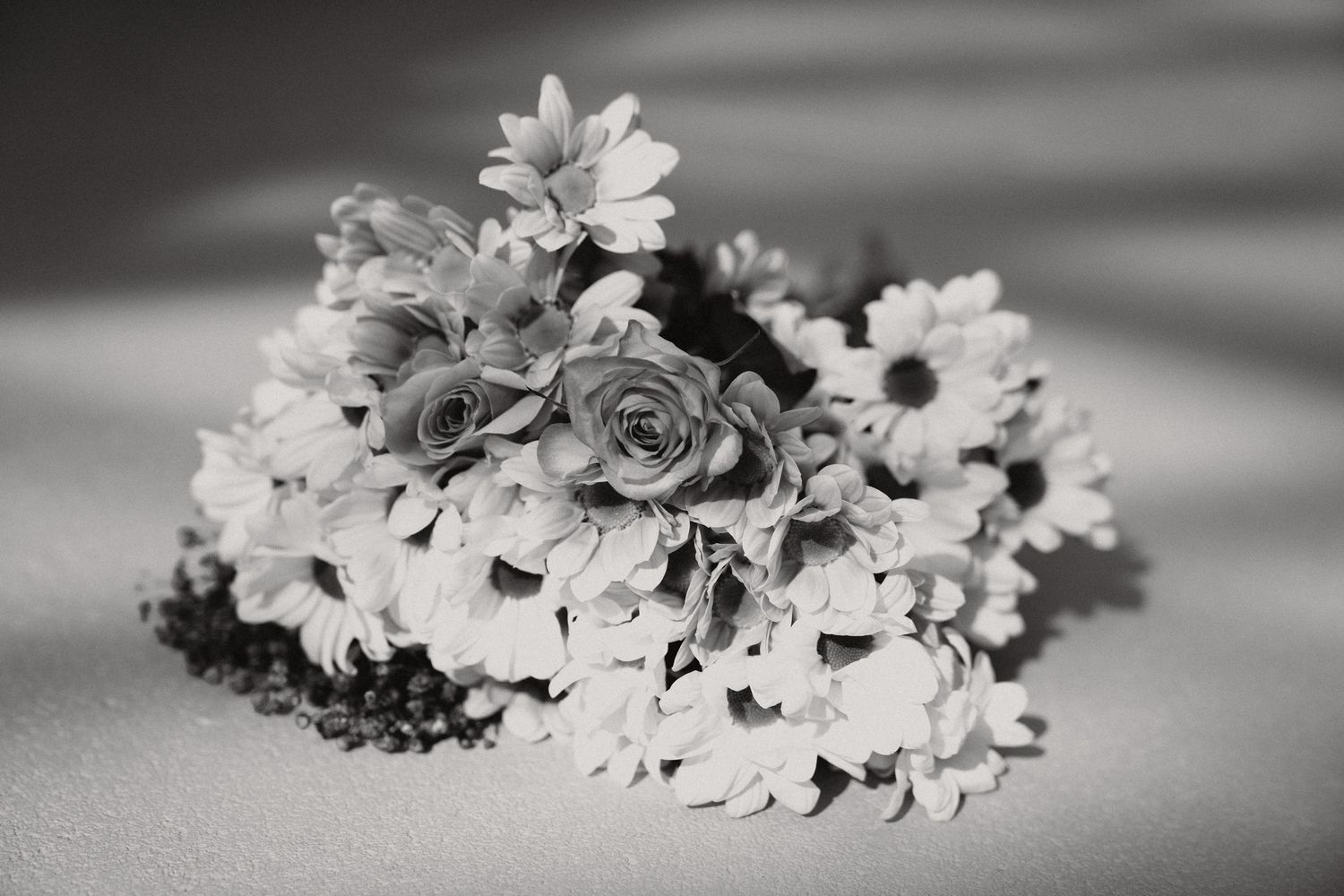 Black and white photo of a bridal bouquet featuring roses and daisies arranged on a soft surface.