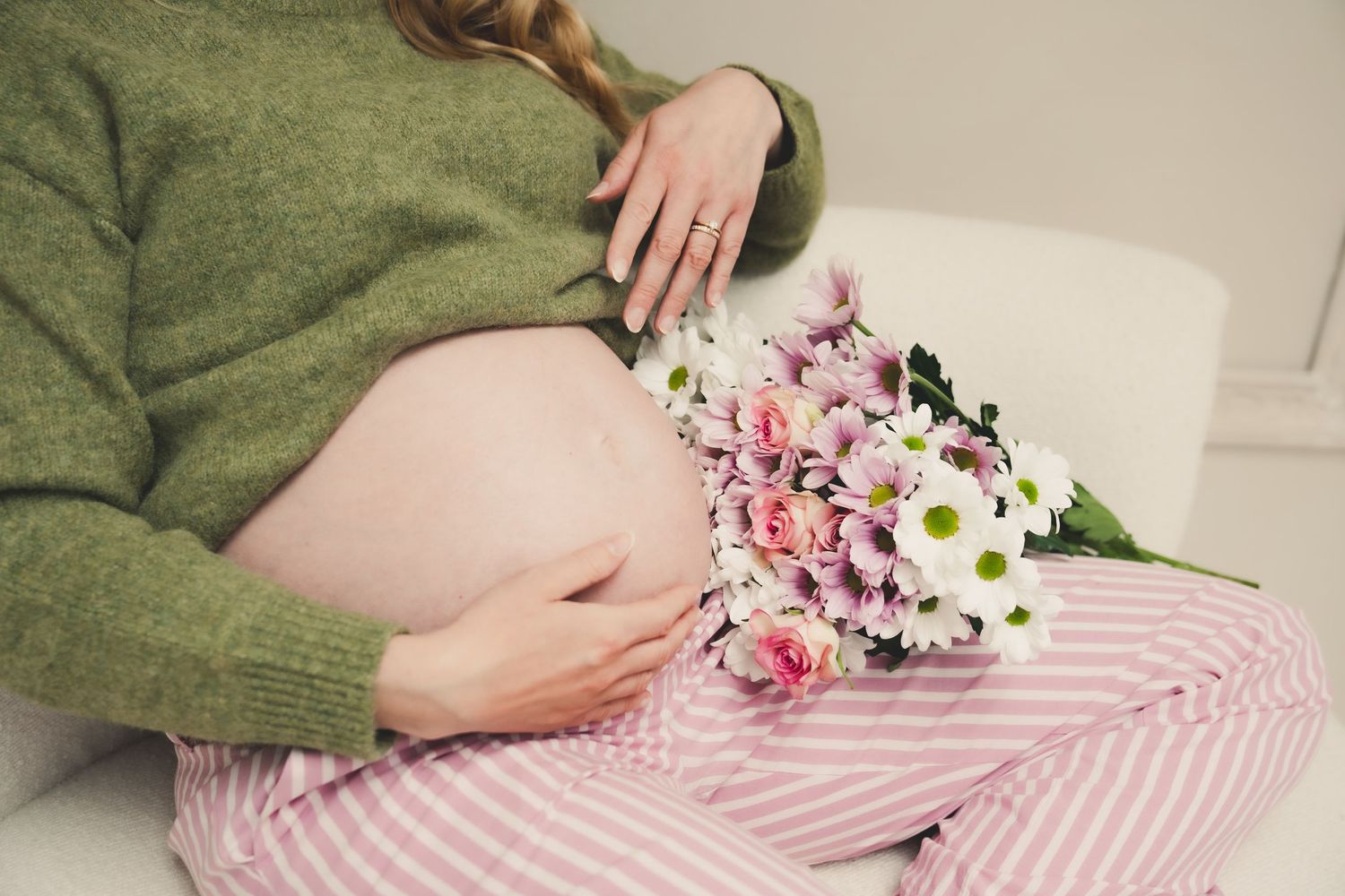 Pregnant woman in green sweater and pink striped pants holding a bouquet of pink and white flowers while cradling her baby bump.