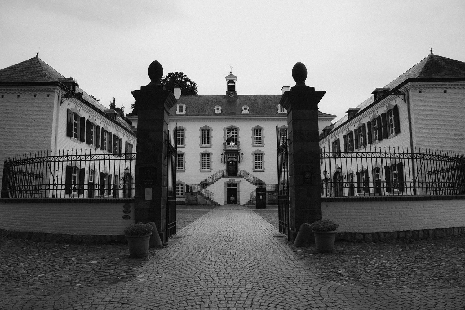 Black and white photo of a grand historic manor house with ornate iron gates and cobblestone pathway.