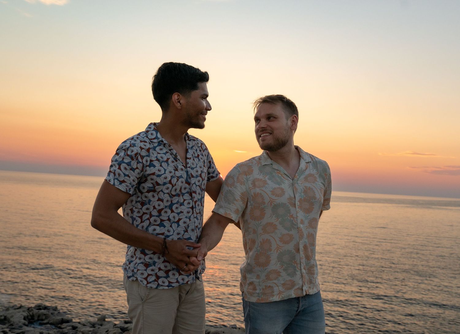 Two men in floral shirts smile at each other by the ocean during a colorful sunset.
