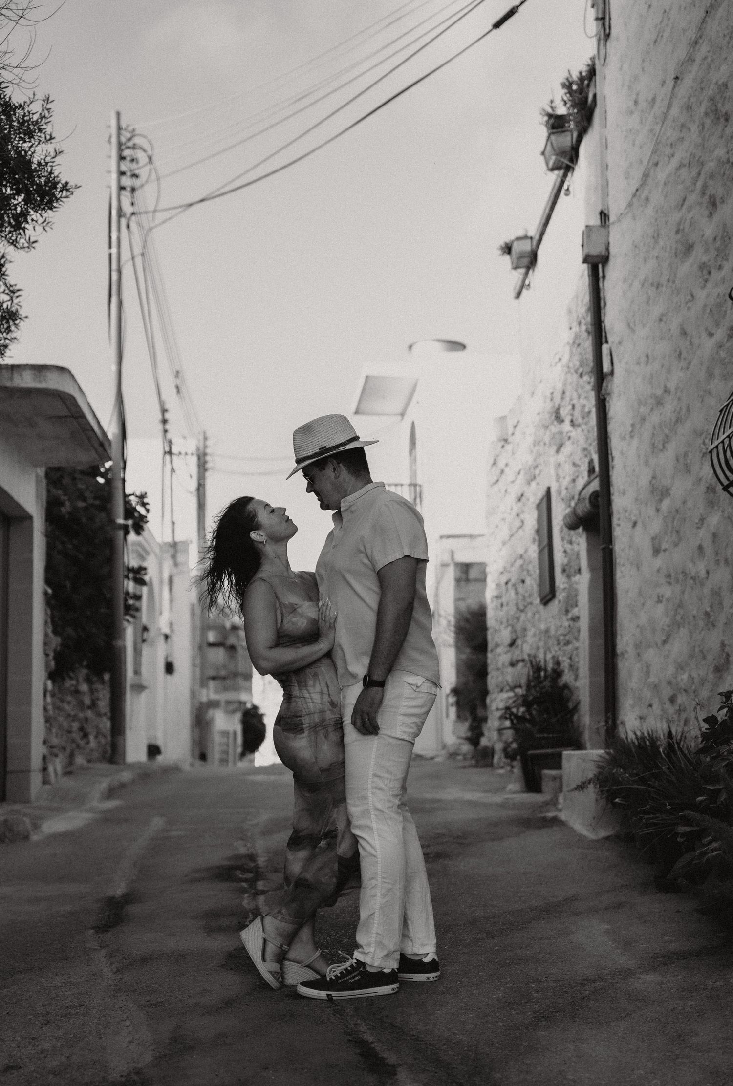 Black and white photo of a romantic couple almost kissing in a narrow Mediterranean alley, man wearing a white hat.