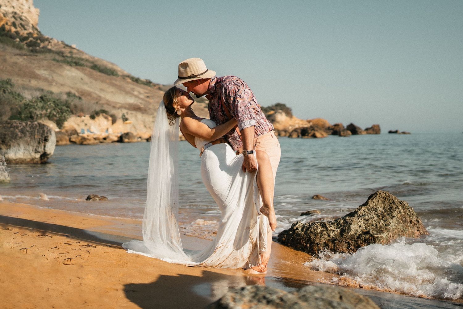 A romantic beach wedding photo with couple embracing near ocean rocks at golden hour.
