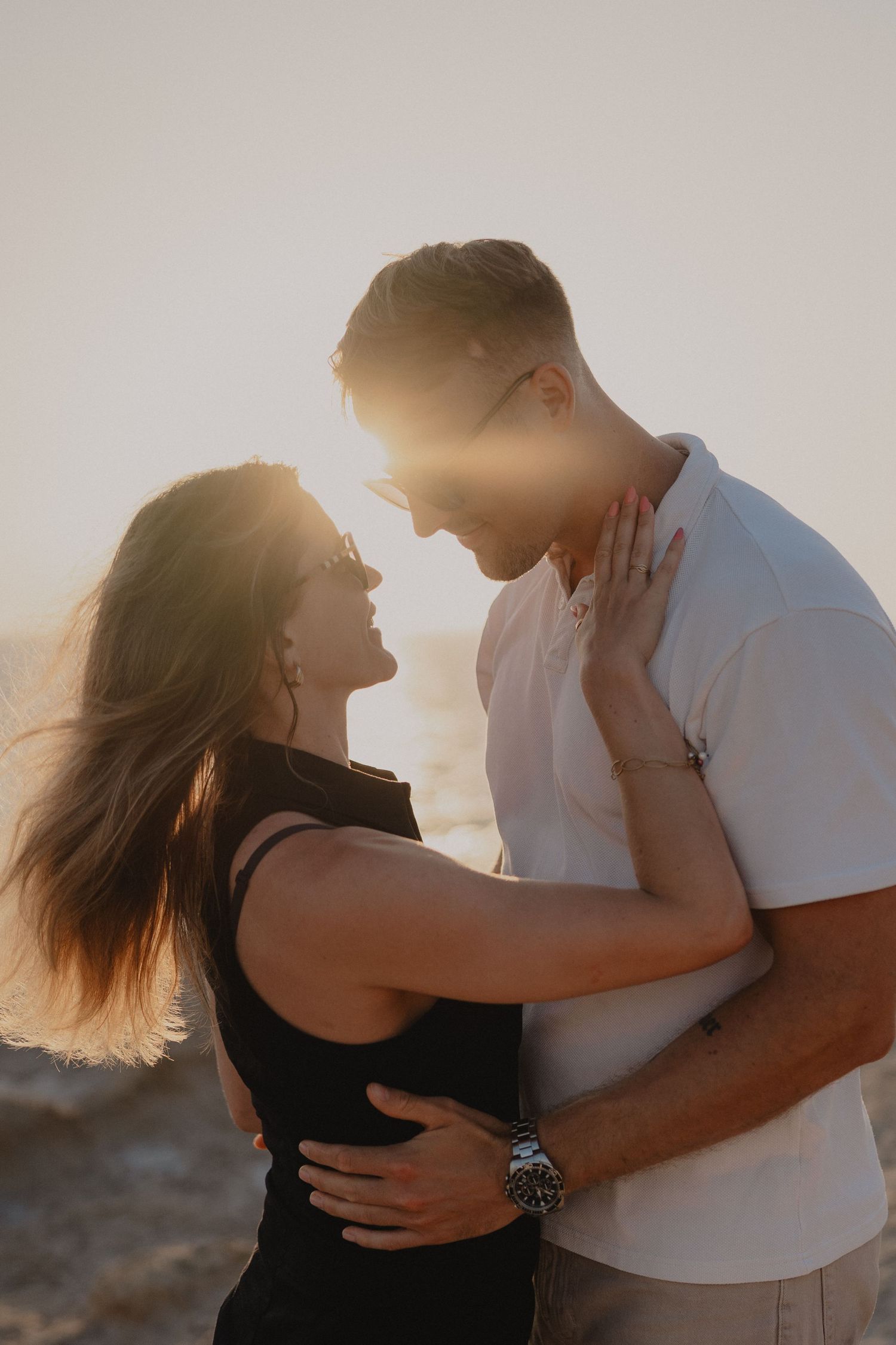 Couple embracing at golden hour sunset, woman in black top and man in white shirt sharing a romantic moment outdoors.