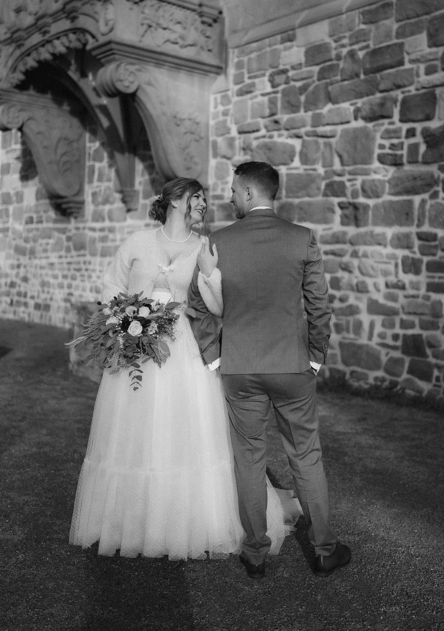 Black and white photo of newlyweds walking along a stone wall in elegant wedding attire.