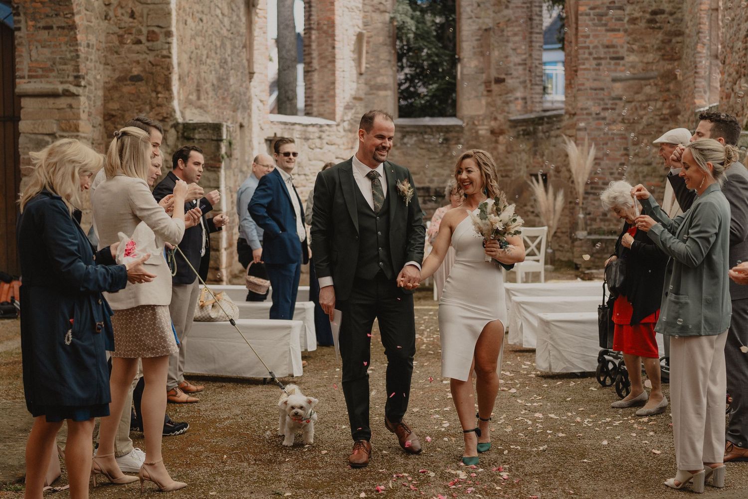 Wedding guests throw confetti at a couple outside a rustic stone building during a celebration.