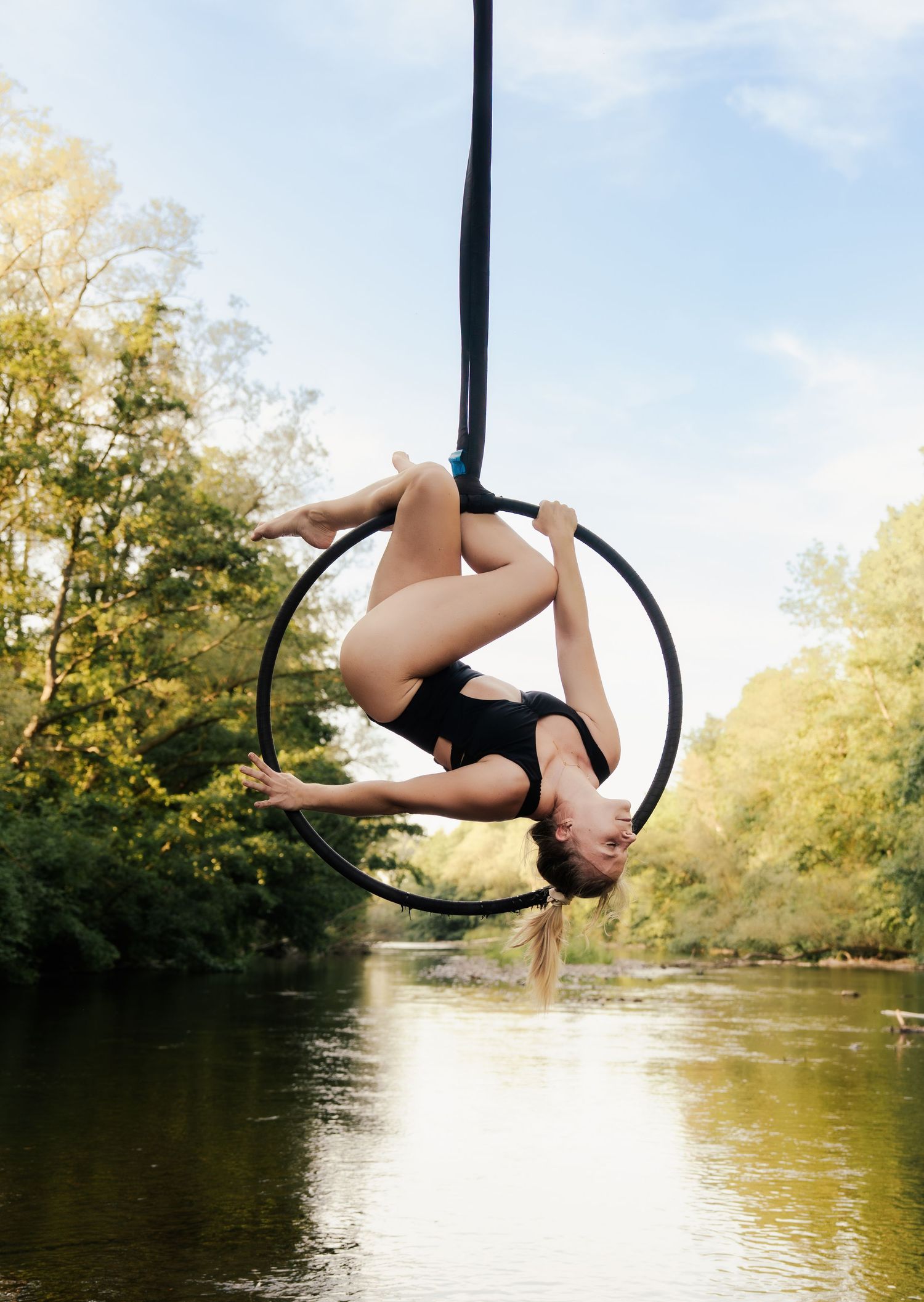 Aerial hoop performer in black leotard gracefully balancing on lyra over a serene river surrounded by lush green trees.