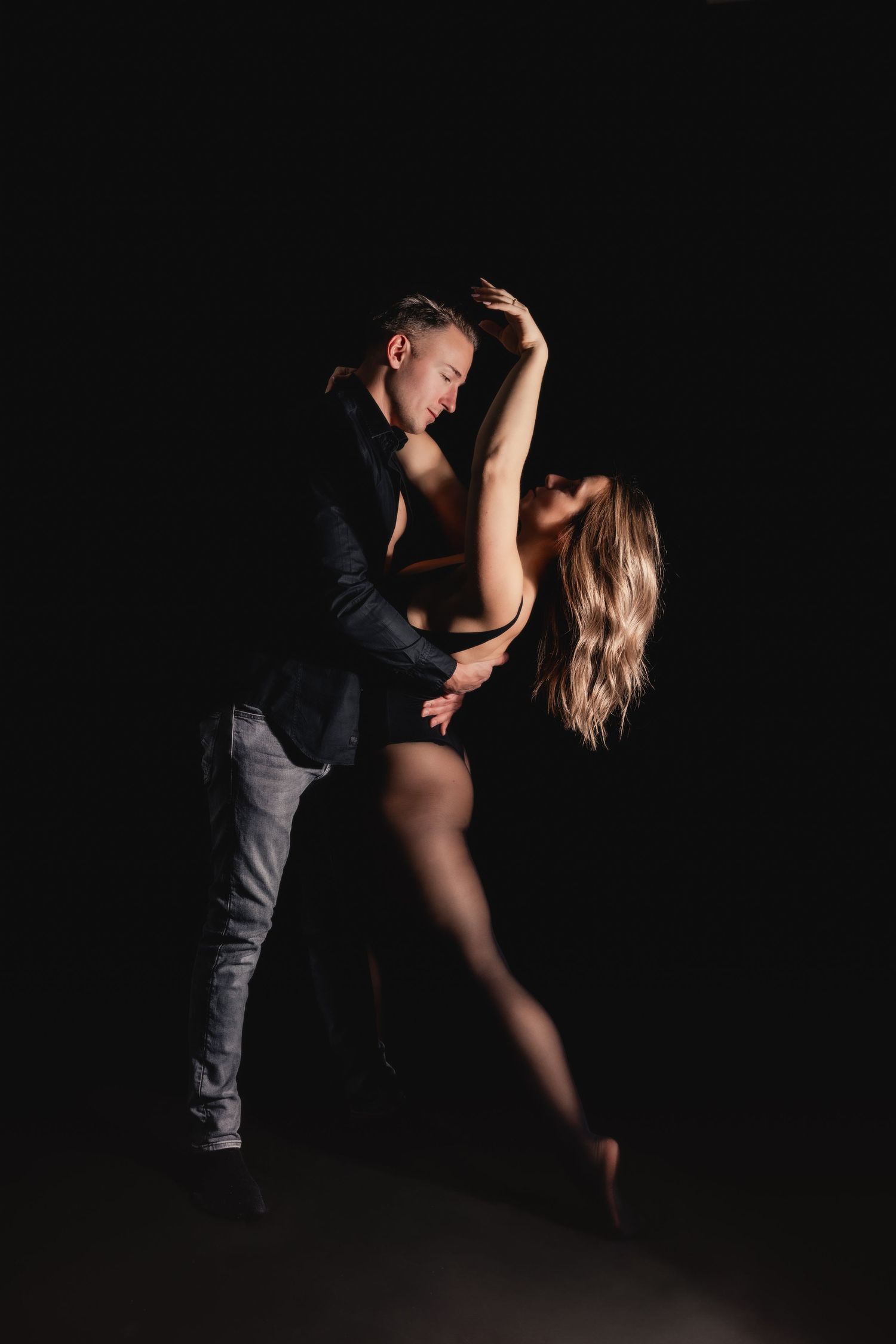 A couple dances intimately in dark studio, woman in bodysuit and stockings, man in blazer and jeans.