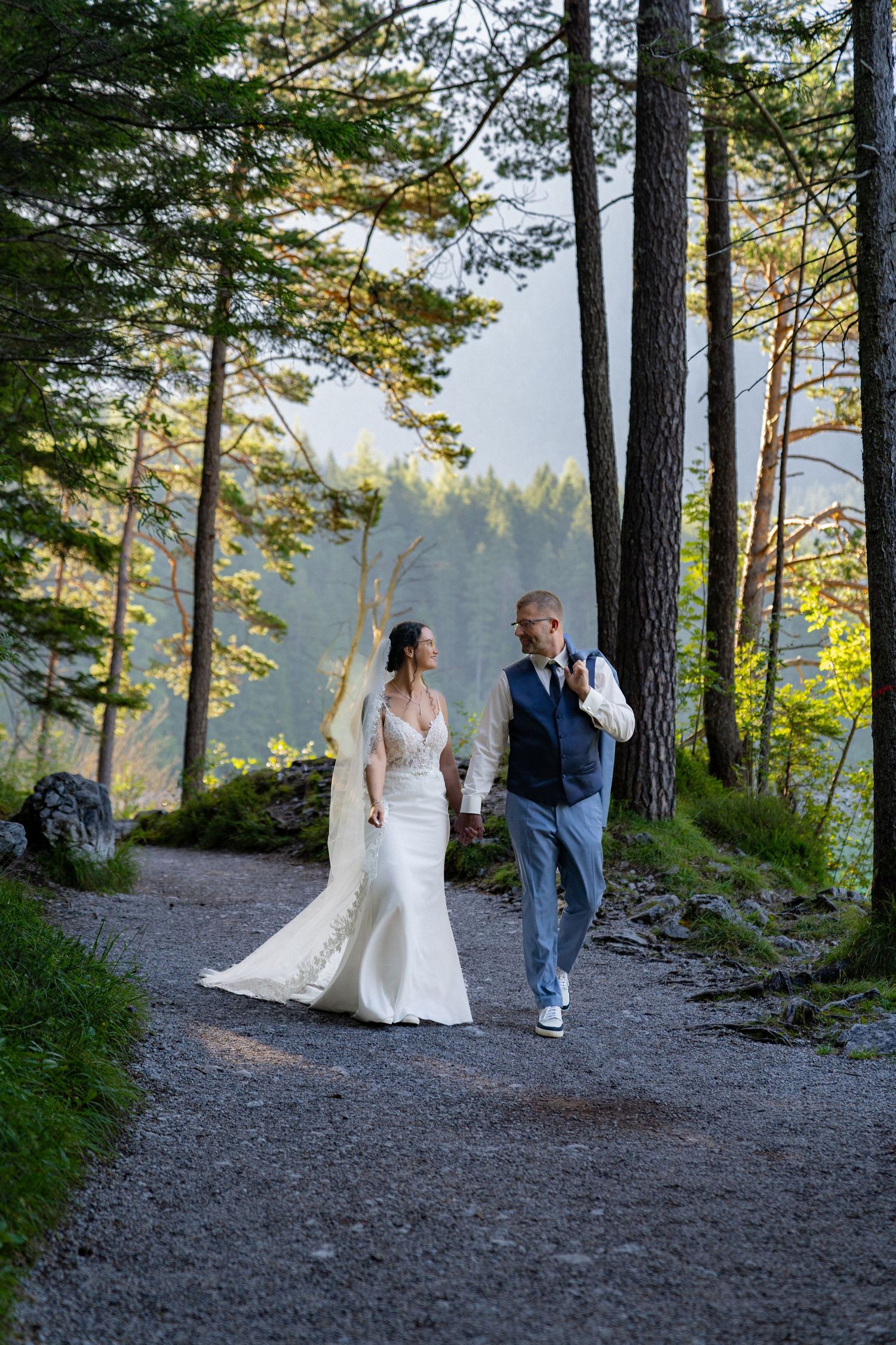 A couple walks along a scenic forest path surrounded by tall pine trees during a wedding photoshoot at sunset.