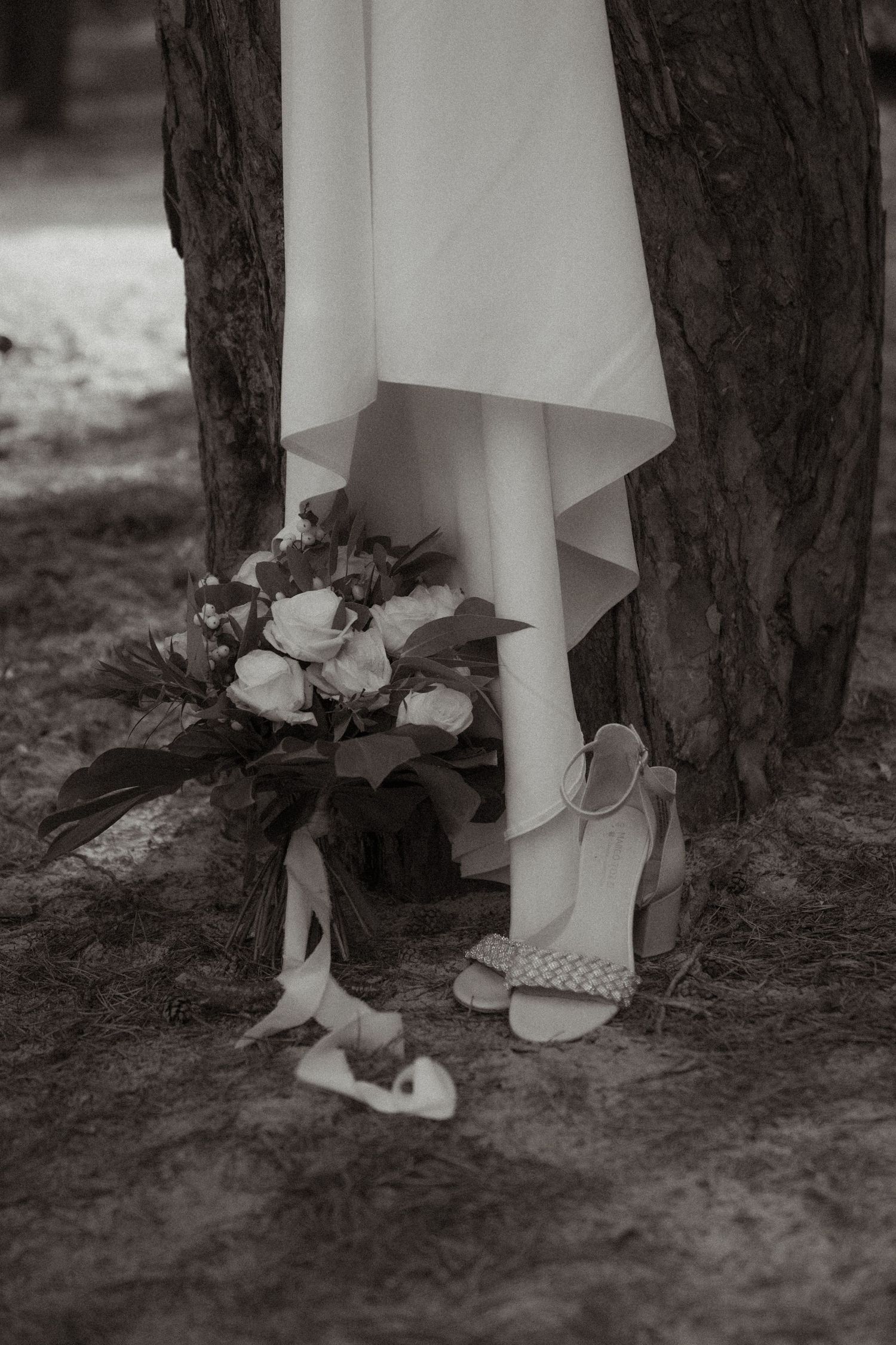 Wedding shoes and bouquet arranged beneath flowing white dress against dark tree trunk.