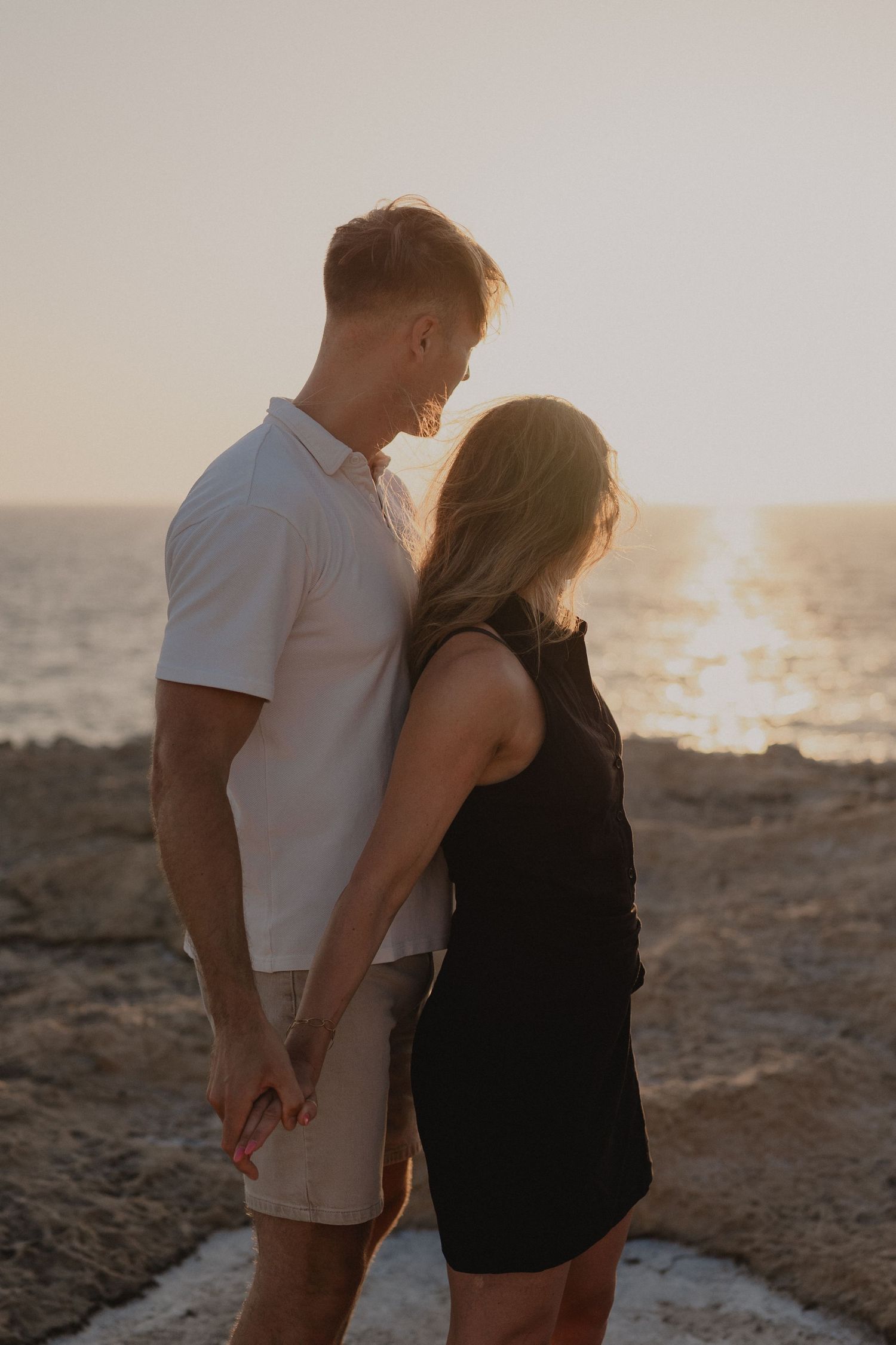 Couple holding hands at sunset by the ocean, man in white shirt and woman in black dress on rocky shore.