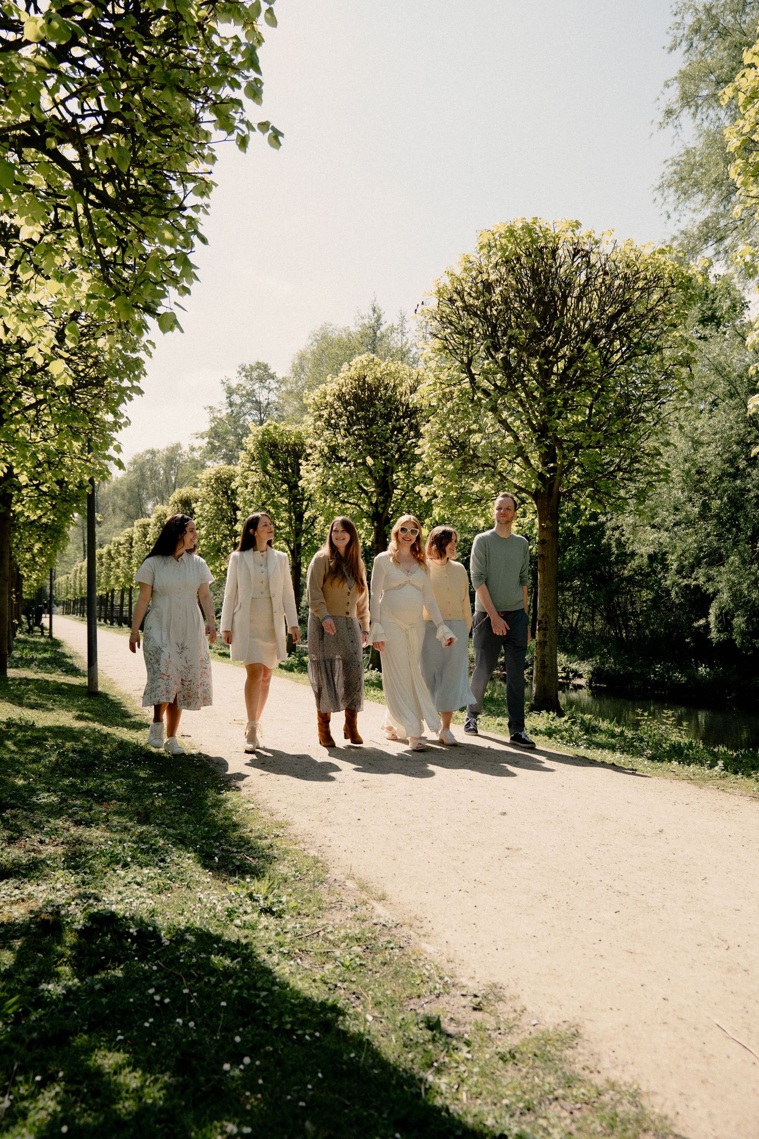 Group of five people strolling along a scenic tree-lined park path on a bright sunny day.