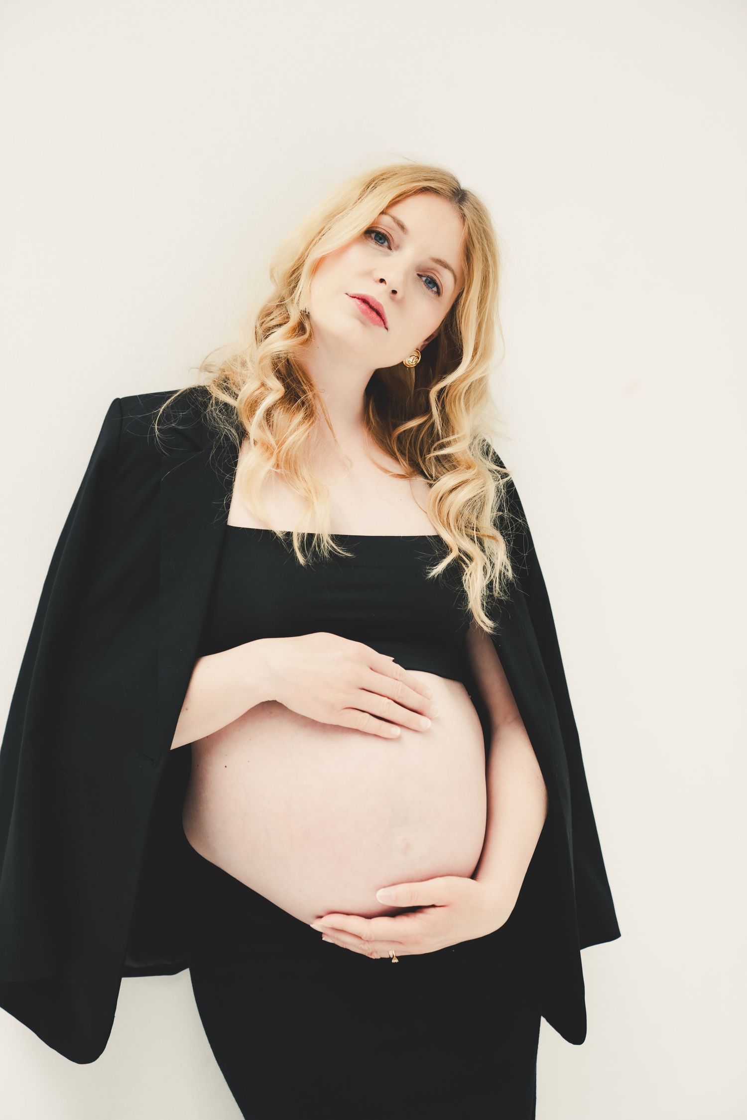 Pregnant blonde woman in black dress and blazer cradling her baby bump against a white background.