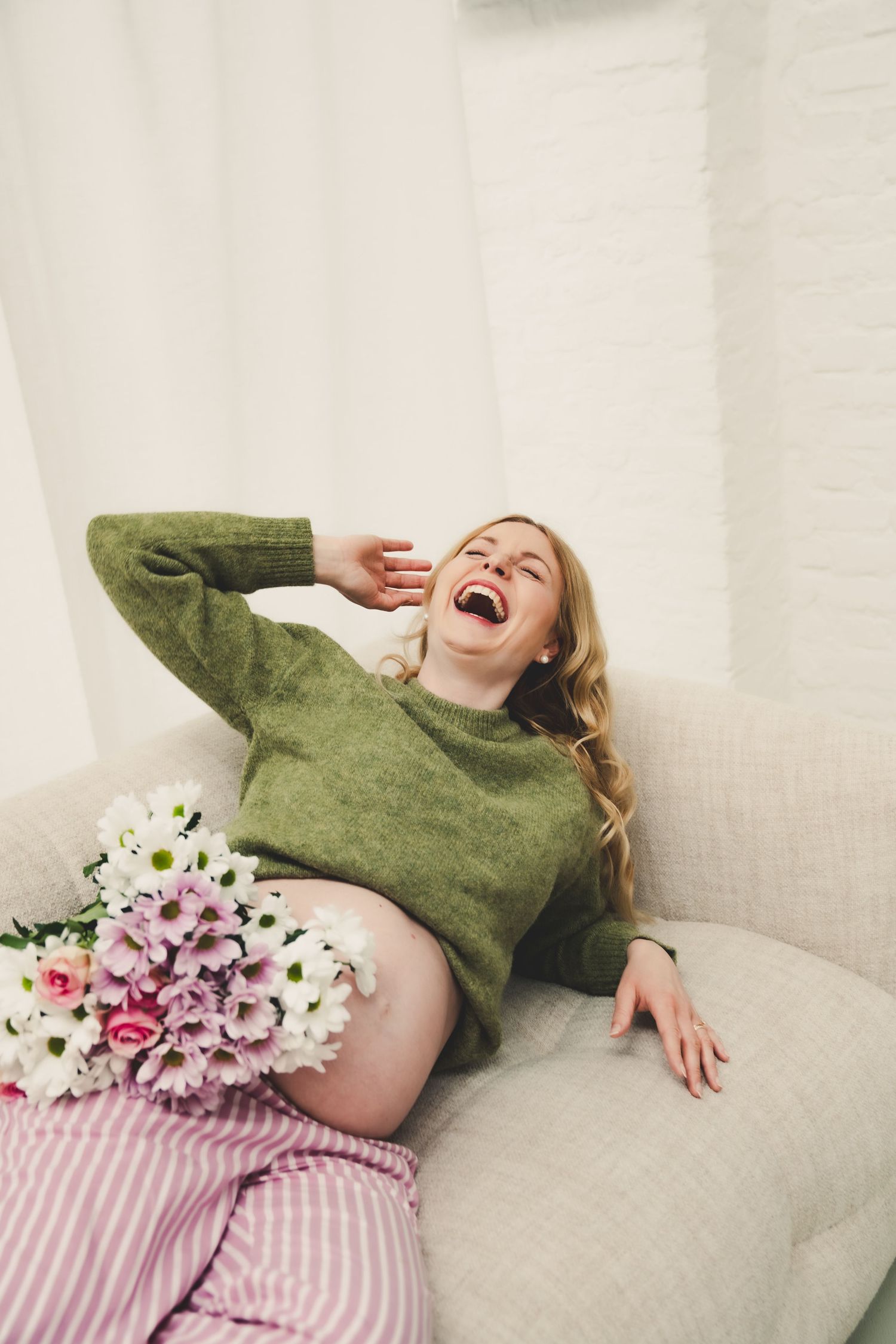 Laughing pregnant woman in green sweater relaxing on sofa with bouquet of pink and white flowers.