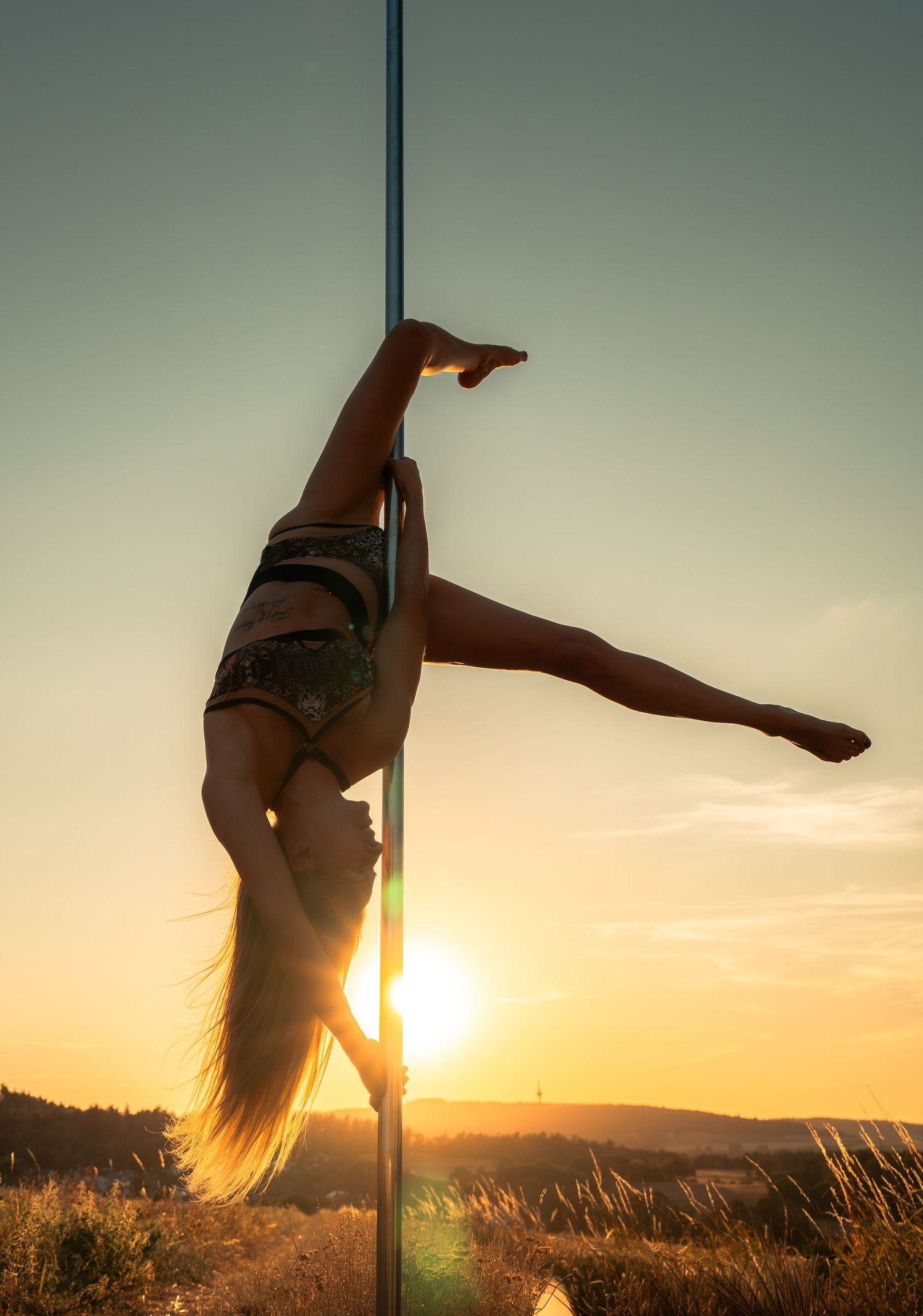 Woman performing pole dance outdoors at sunset, elegantly inverted with legs extended against golden sky.