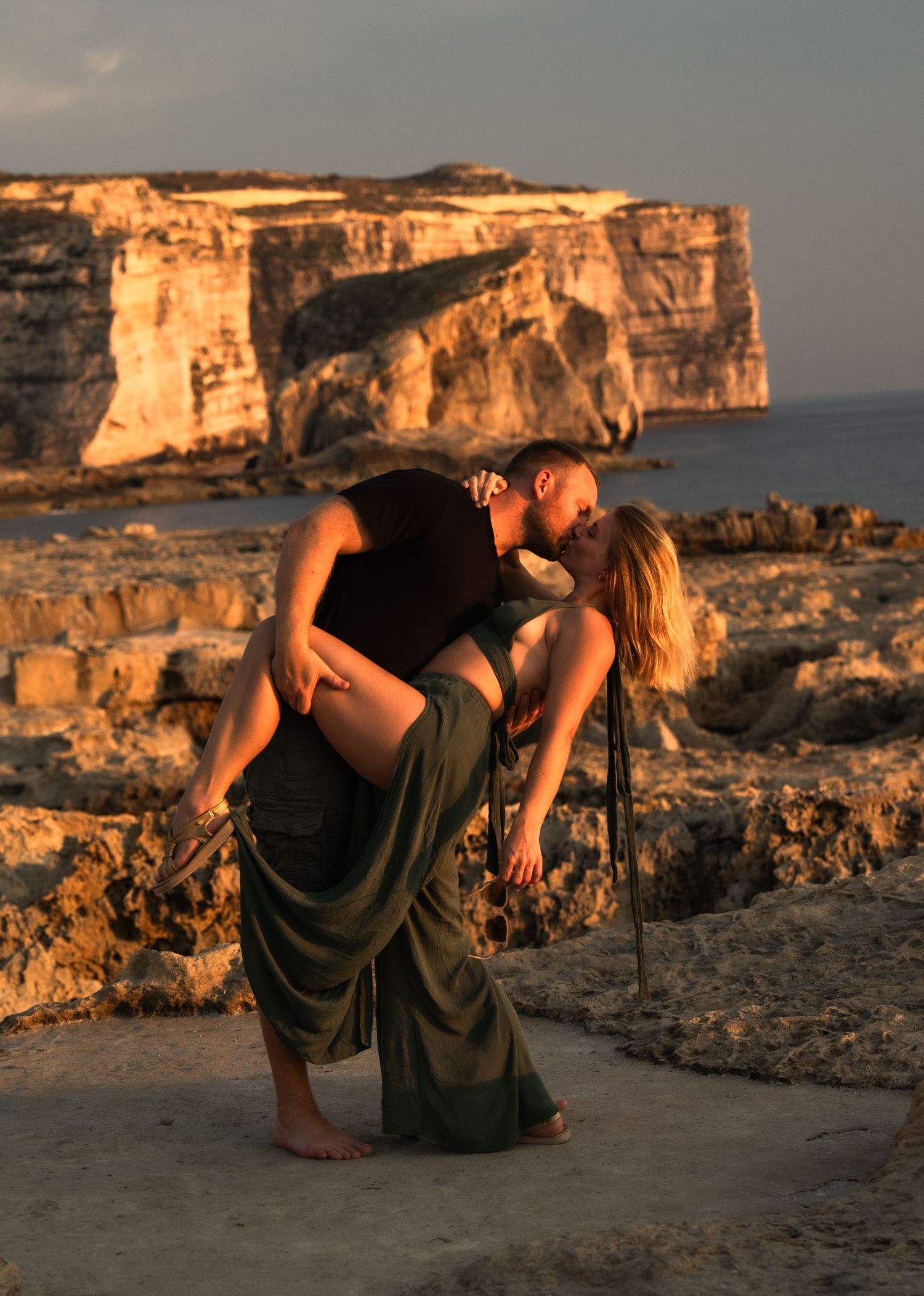 Couple sharing a romantic dip kiss at golden hour on rocky coastal cliffs with ocean backdrop.