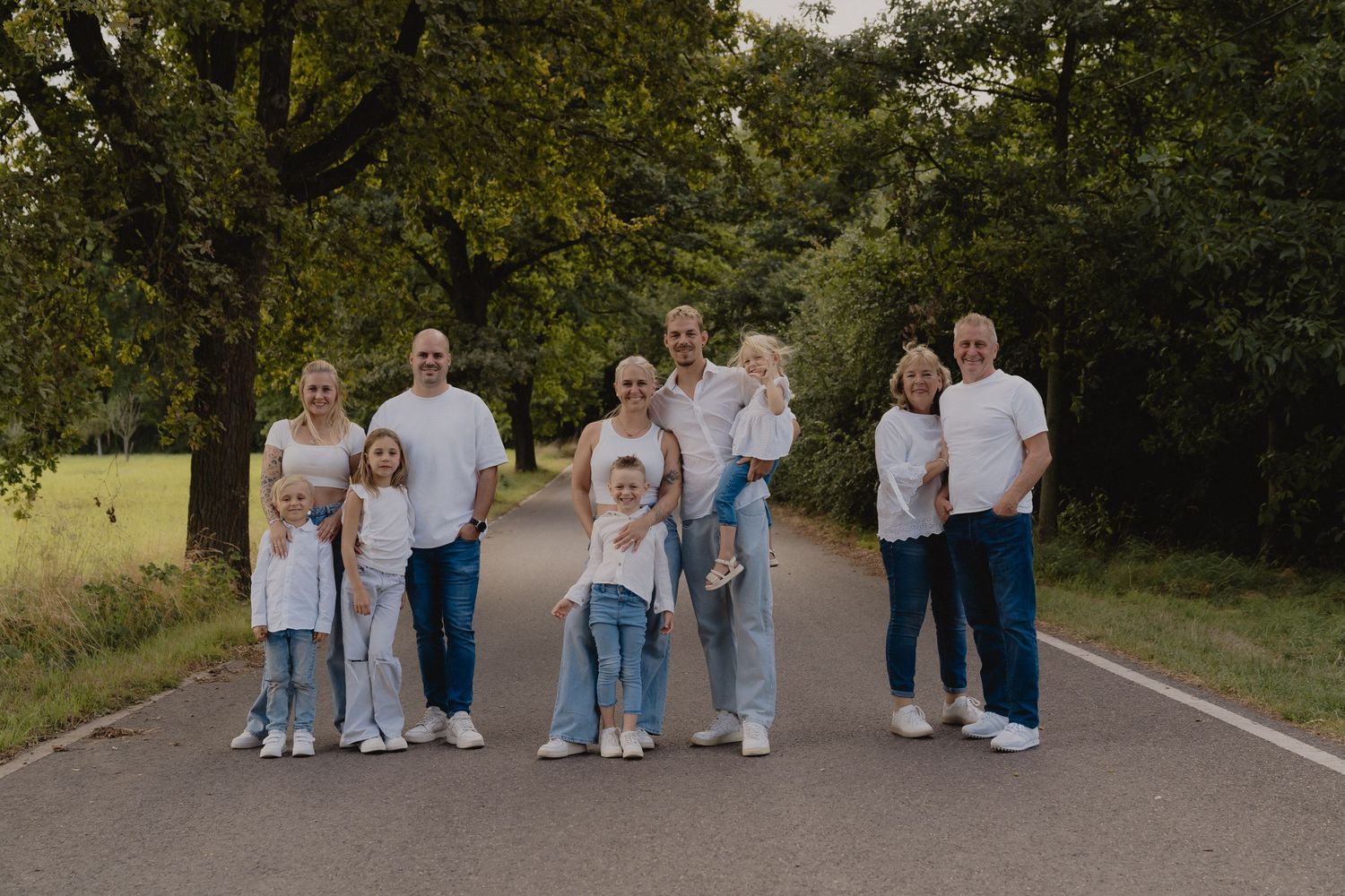 Multi-generational family portrait on a tree-lined path, wearing coordinated white tops and blue jeans outdoors.