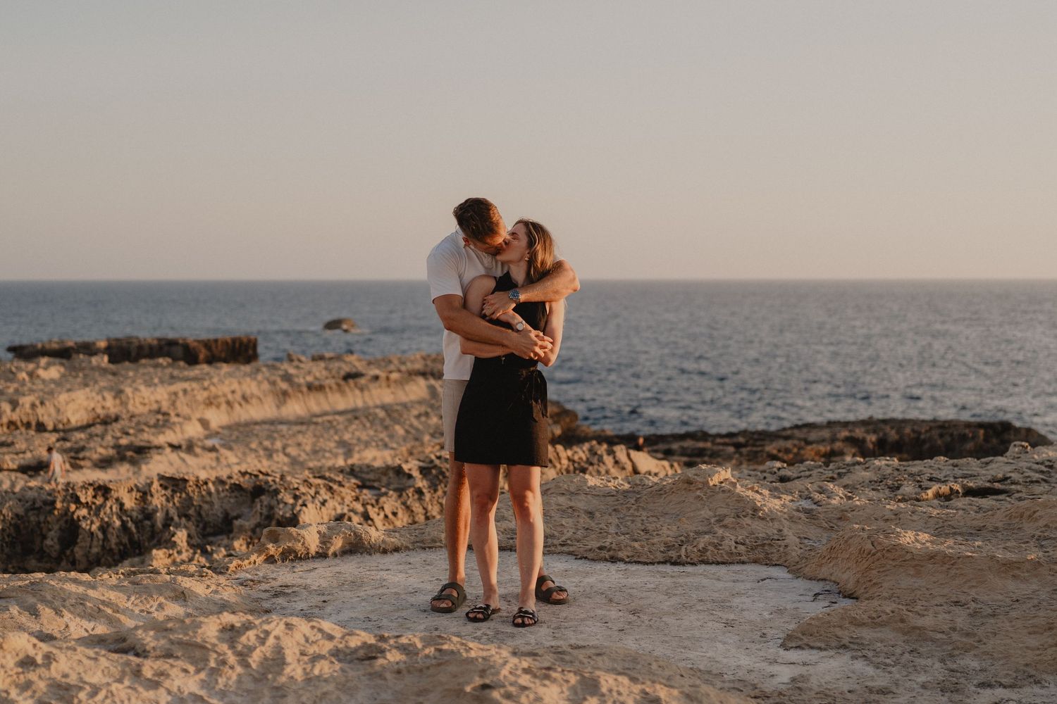 Couple embracing and kissing on rocky coastal cliffs at sunset with calm ocean in the background.