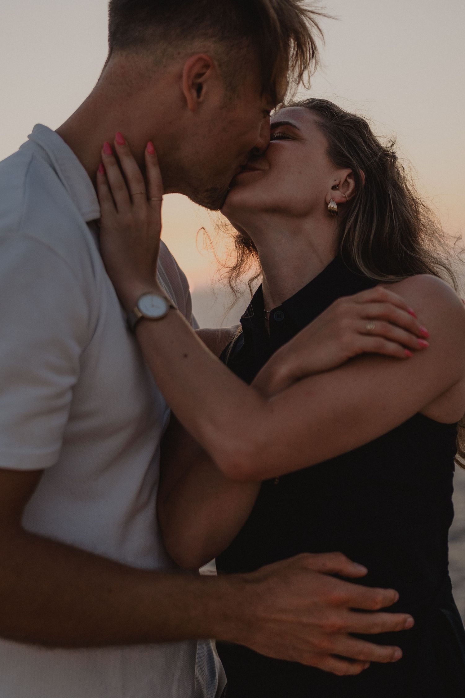 Romantic couple sharing a passionate kiss at sunset, man in white shirt and woman in black dress embracing outdoors.