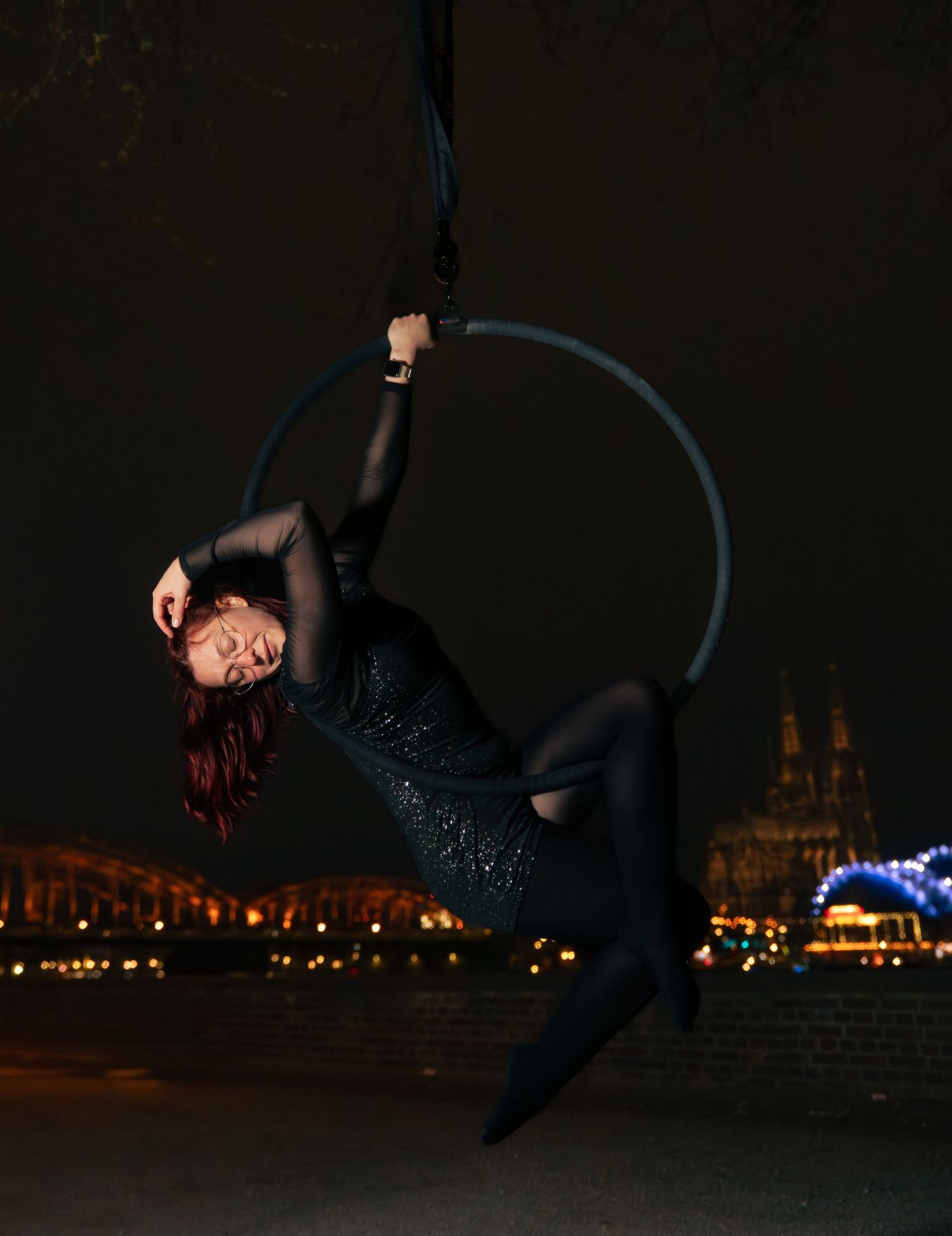 Aerial hoop performer in black outfit poses gracefully on lyra at night with city lights in background.