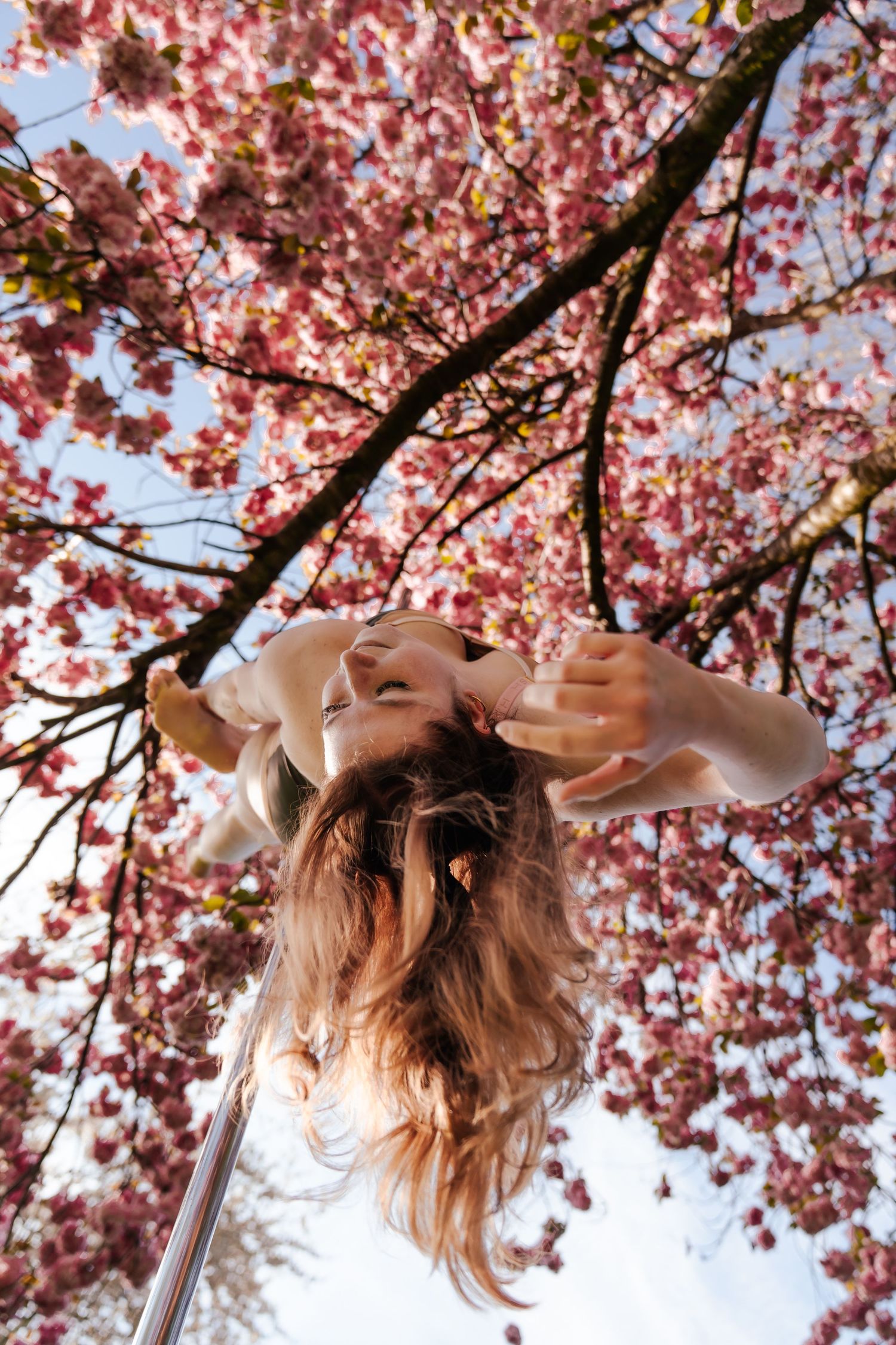 Woman photographed from below, hair flowing down, framed by pink cherry blossom tree branches against a bright sky.