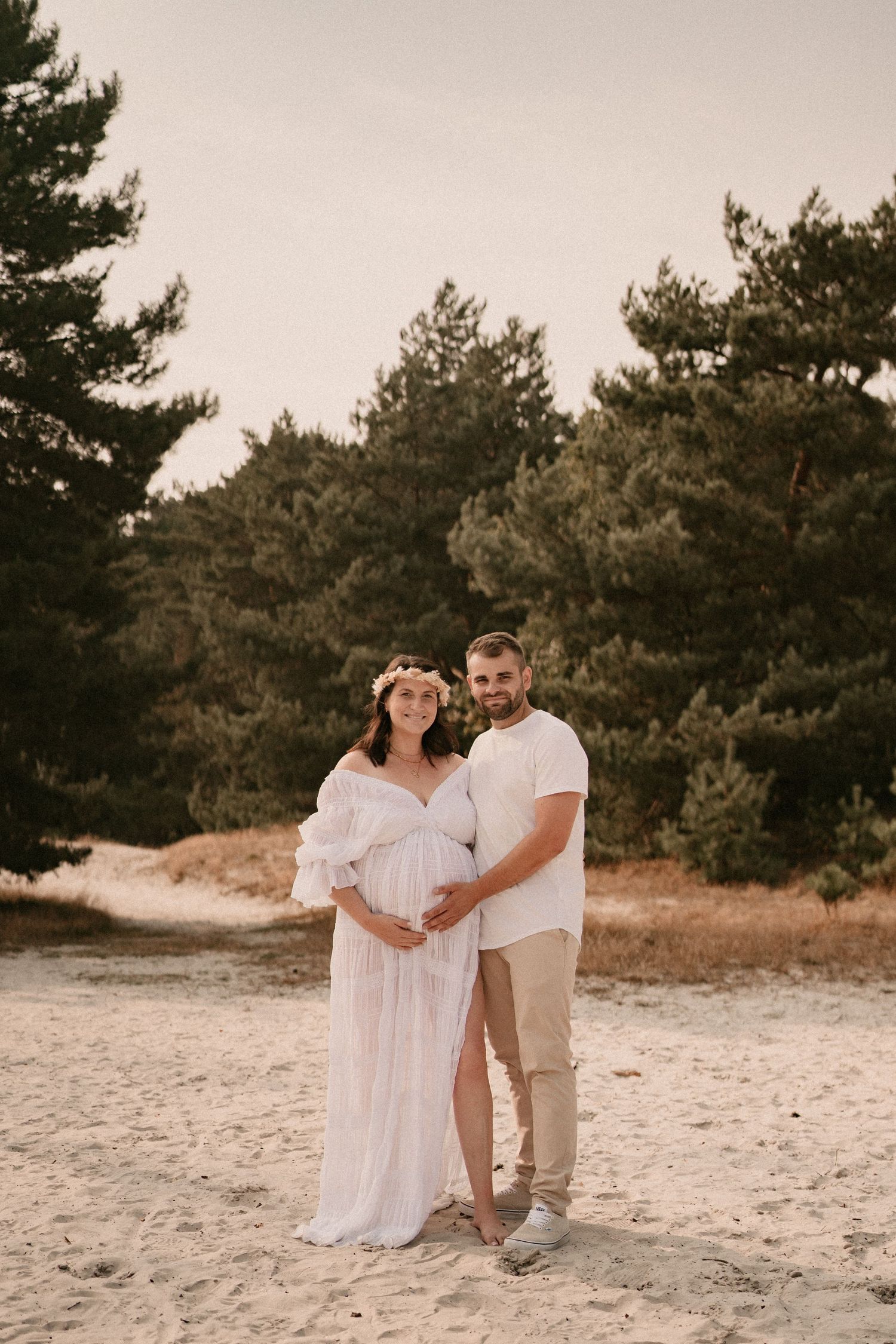 A couple in coordinated white and beige outfits embrace on a sandy beach surrounded by pine trees.