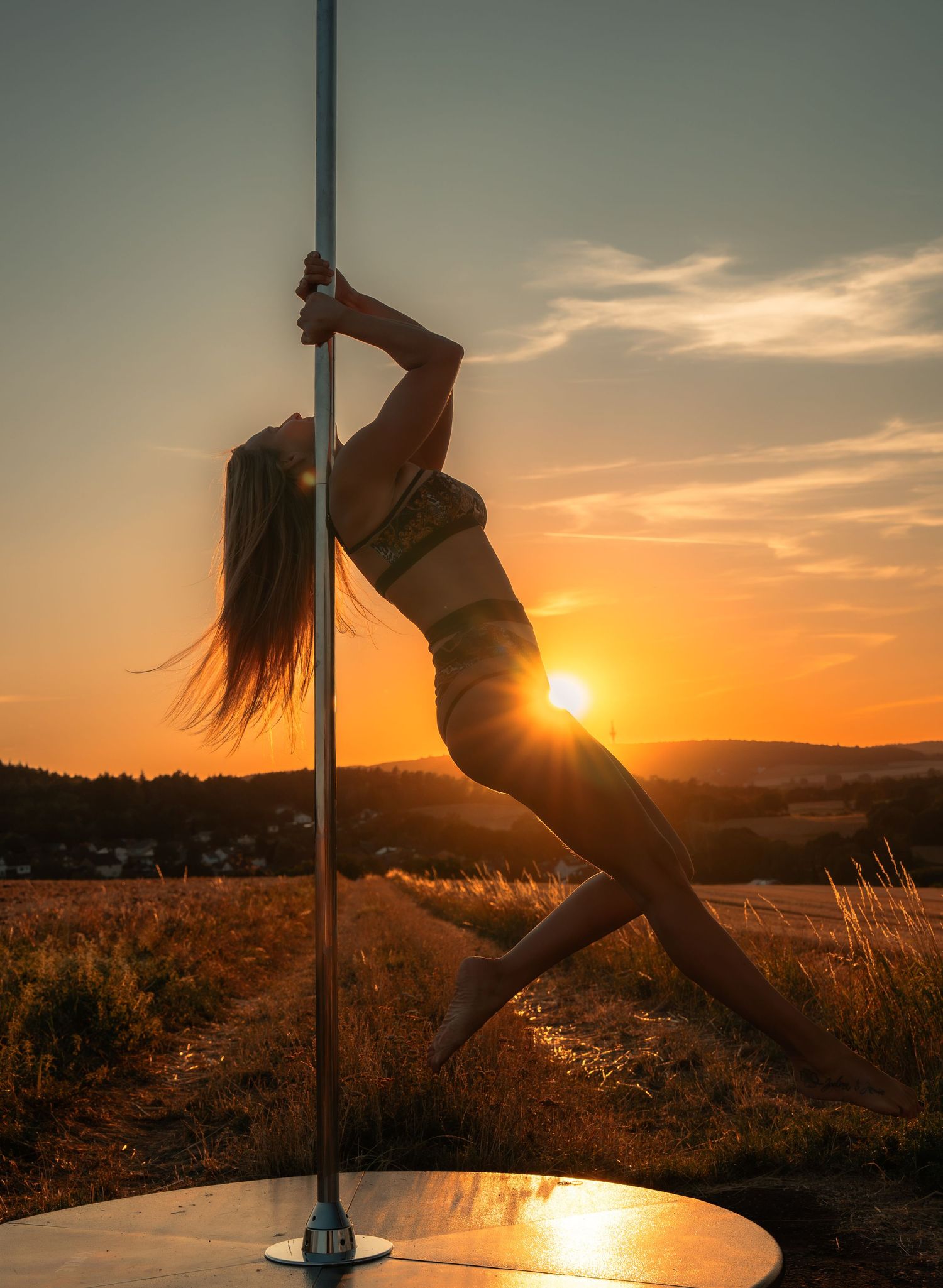 Woman performing pole dance outdoors at golden sunset, showcasing athletic flexibility against a scenic countryside backdrop.