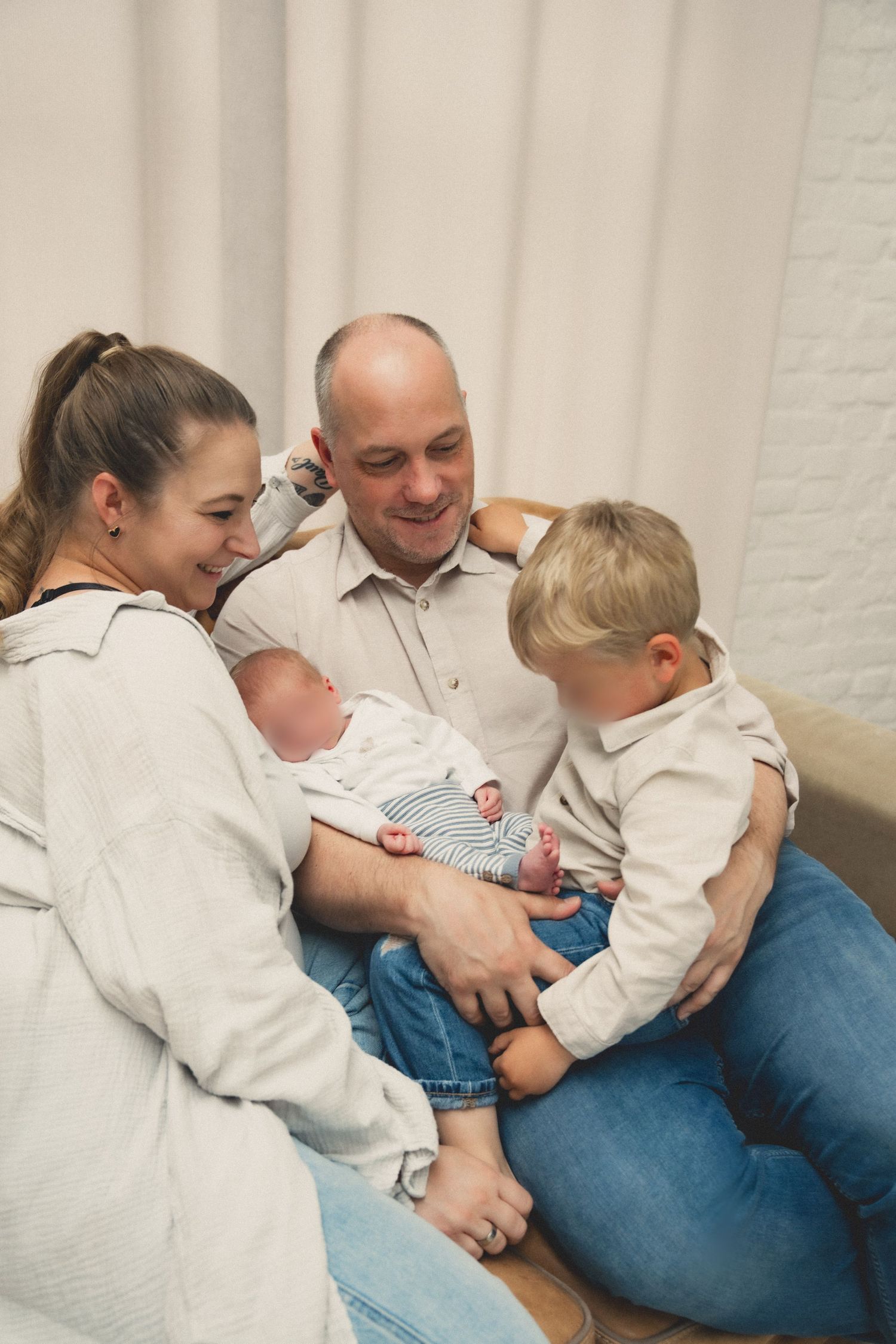 Happy family with parents, toddler, and newborn baby sitting together on a couch in a bright room.
