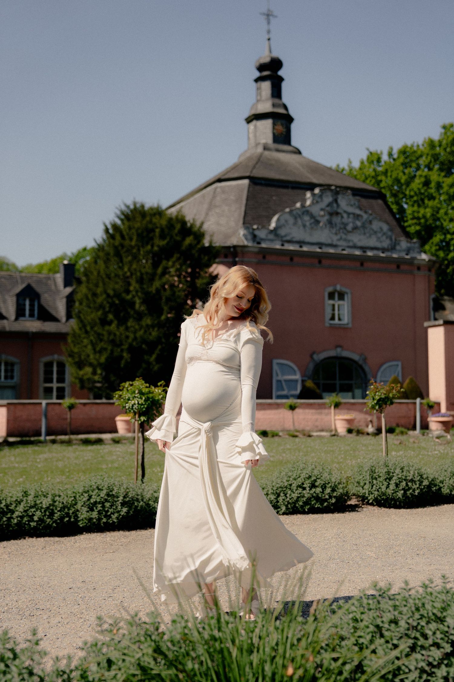 Pregnant woman in elegant white dress poses gracefully in front of a historic pink building on a sunny day.