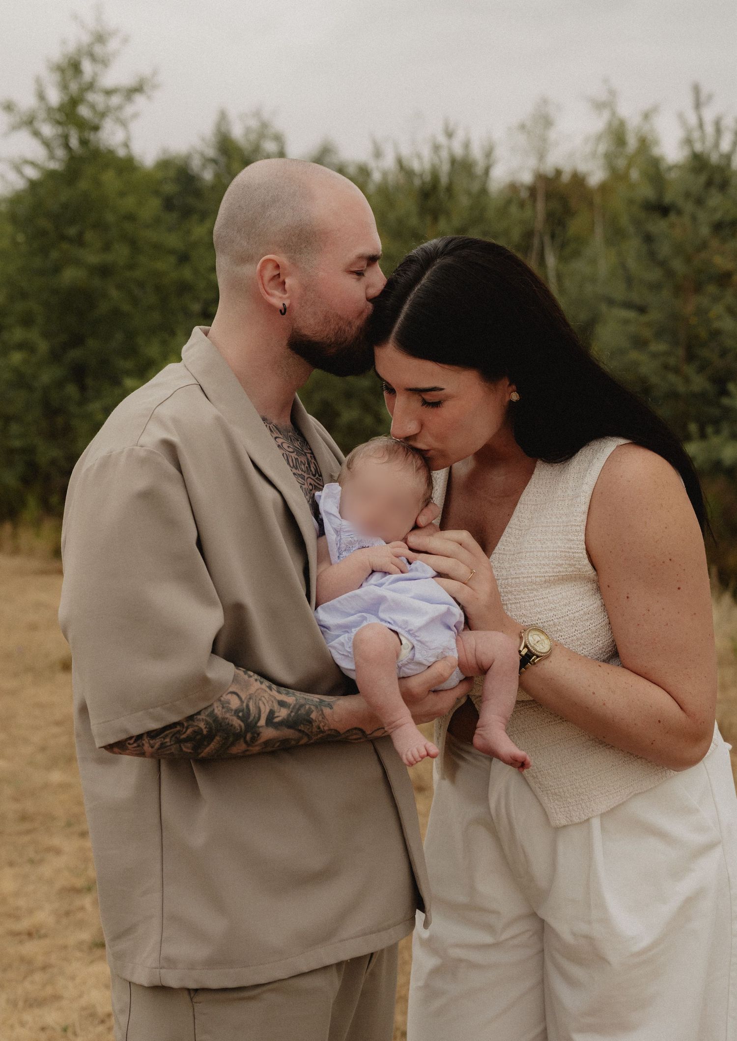 A tattooed man and dark-haired woman share a kiss over their newborn baby outdoors in a field.