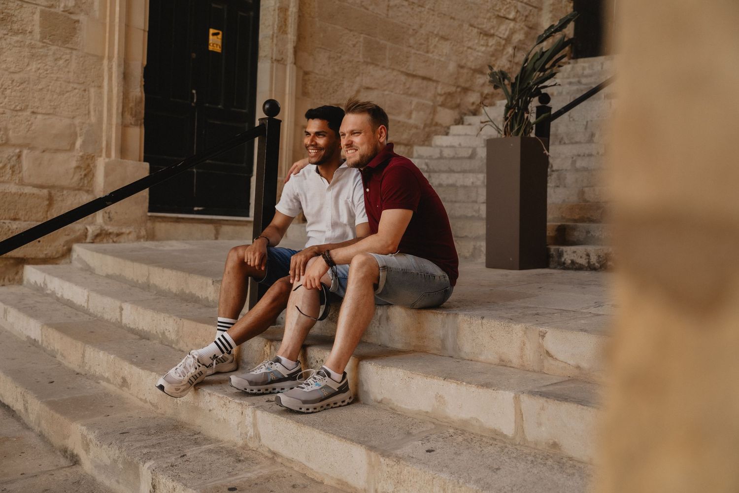 Two men sitting together on stone steps outside a historic building, smiling and relaxing in casual summer outfits.