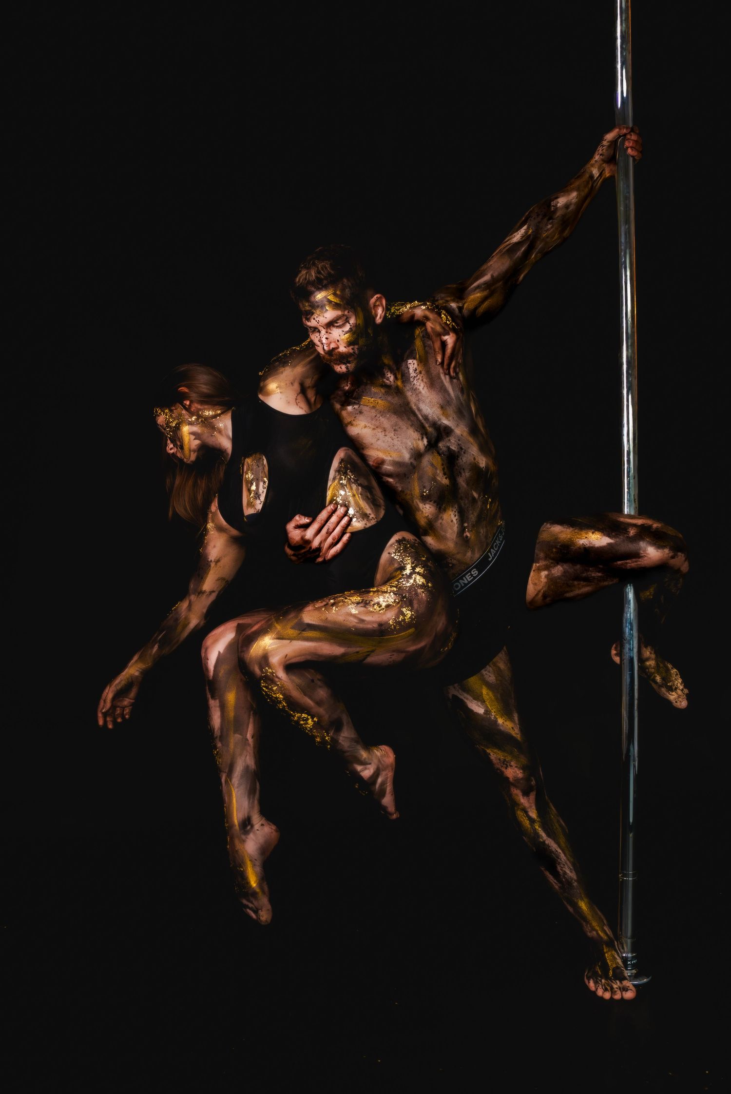 Two body-painted performers with gold artistic makeup pose dynamically on a pole against a black background.