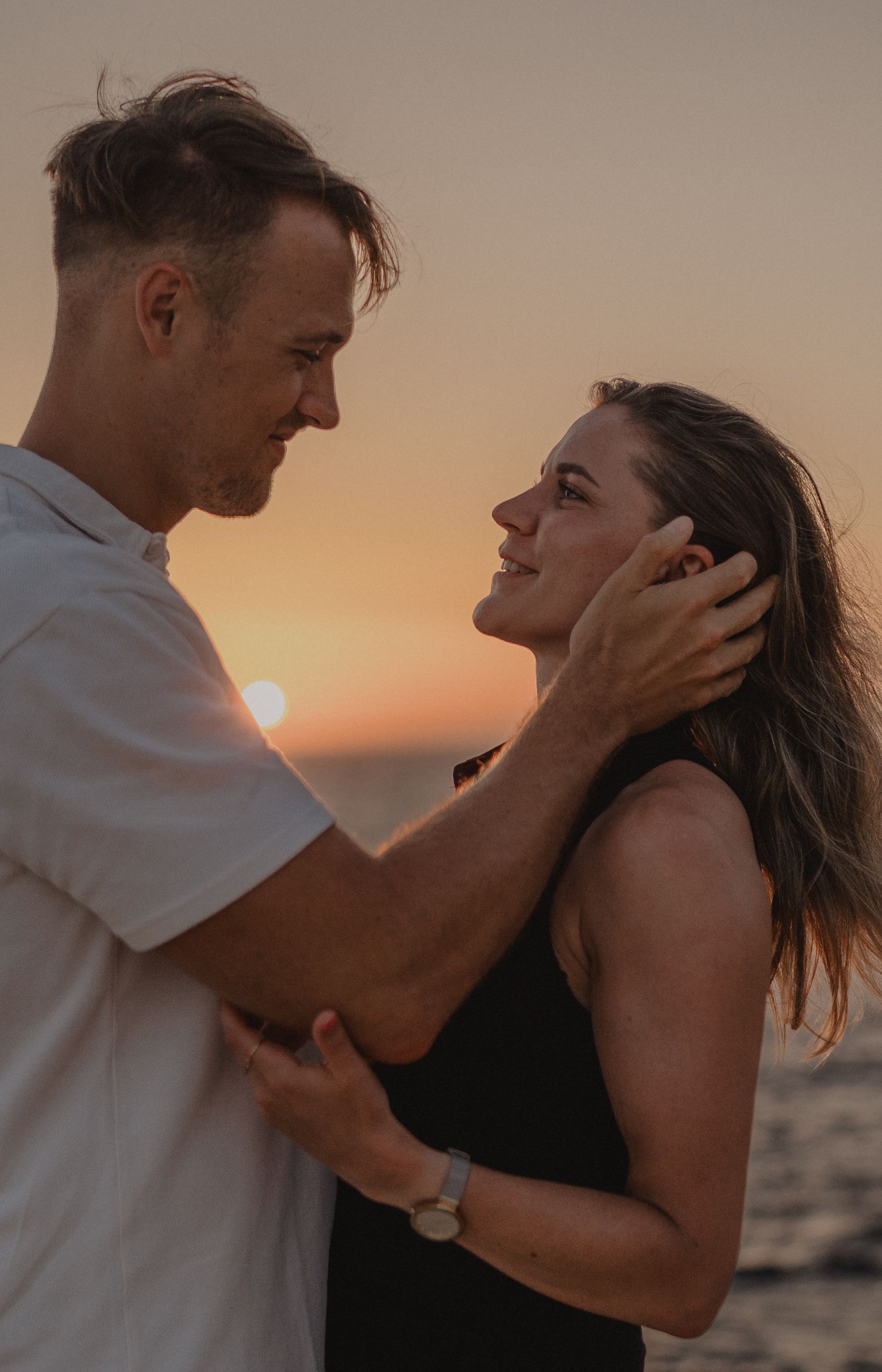 A couple shares a romantic sunset moment on the beach, with the man tenderly holding the woman's face as she smiles up at him.